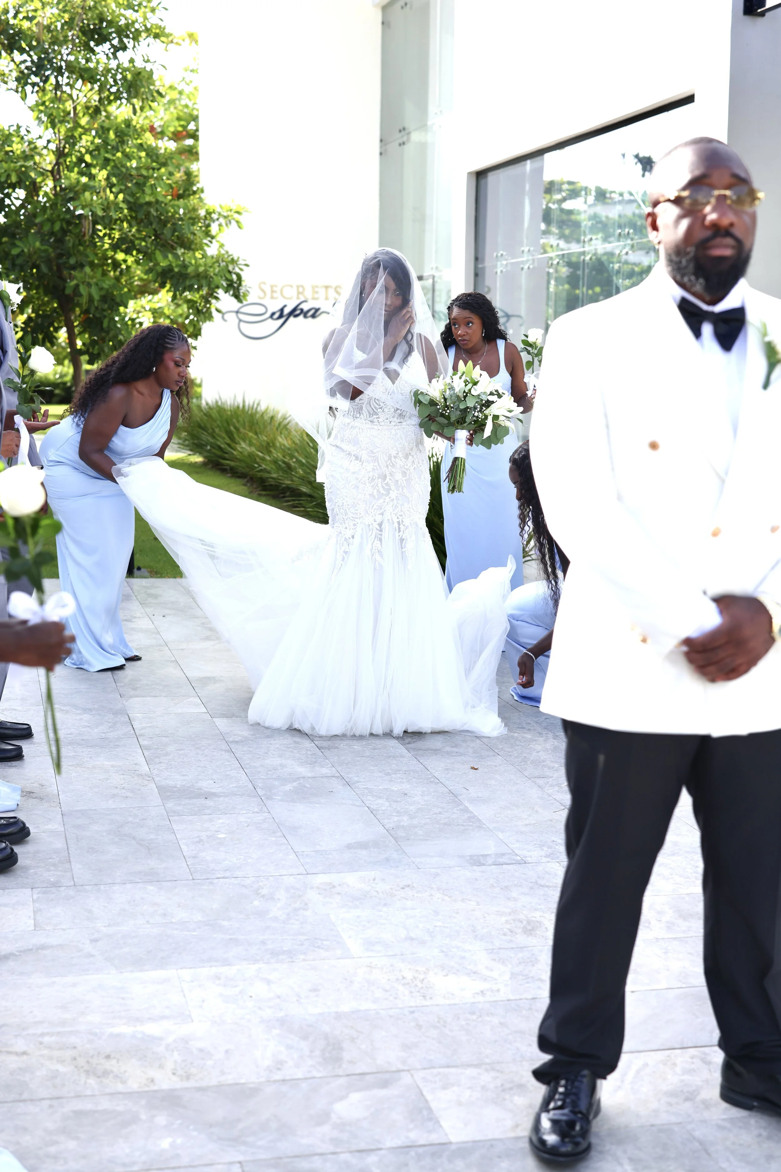 A bride in a white wedding dress and veil holding a bouquet of white flowers stands outside near a modern building and a tree, with bridesmaids in white dresses kneeling and adjusting her train, and a man in a white tuxedo with a black bow tie standi