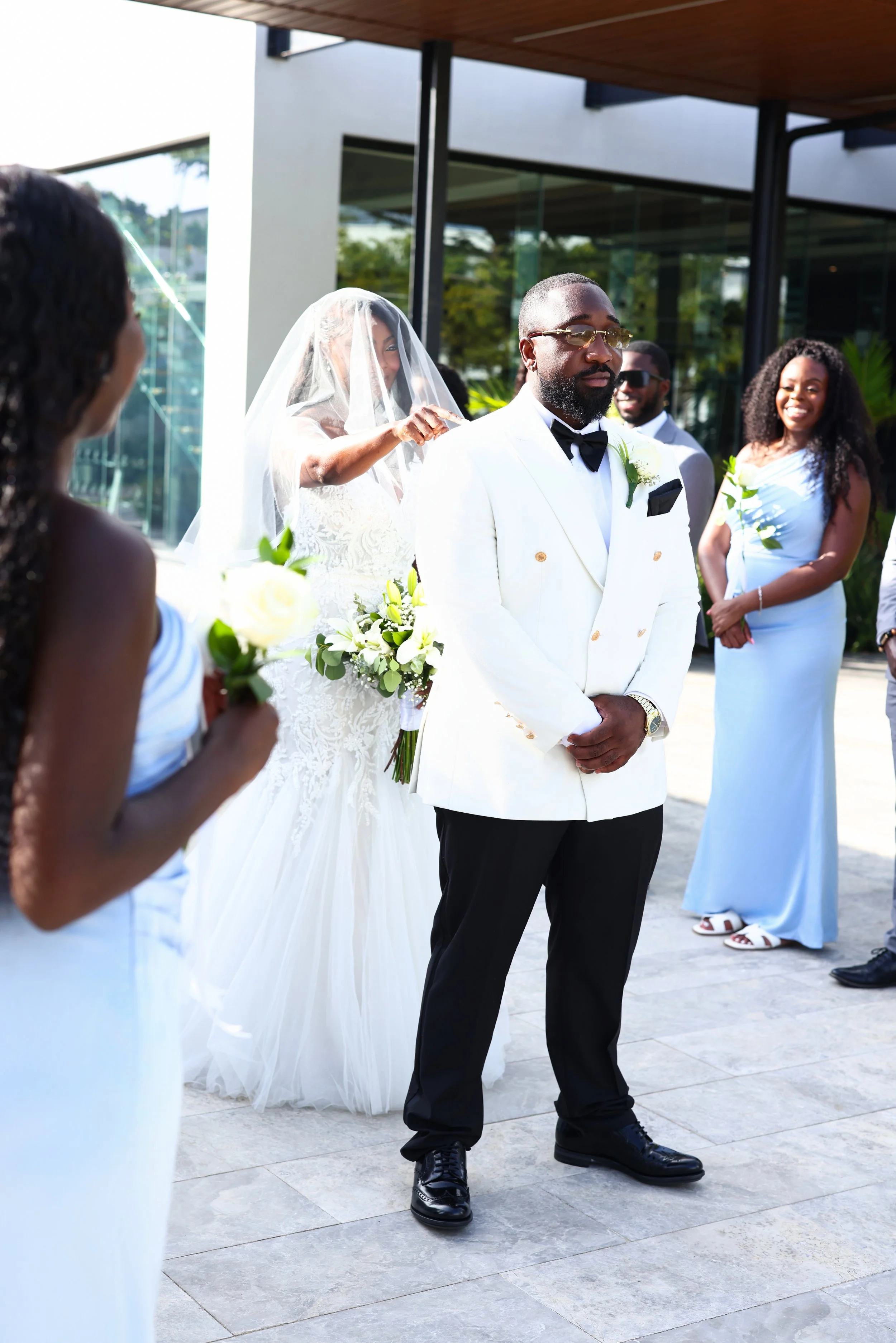 A group of people dressed in wedding attire at a wedding ceremony outside a modern building. The bride is partially visible with a veil, and the groom is in a white suit with a black bow tie, standing with hands clasped. Other guests are smiling and 