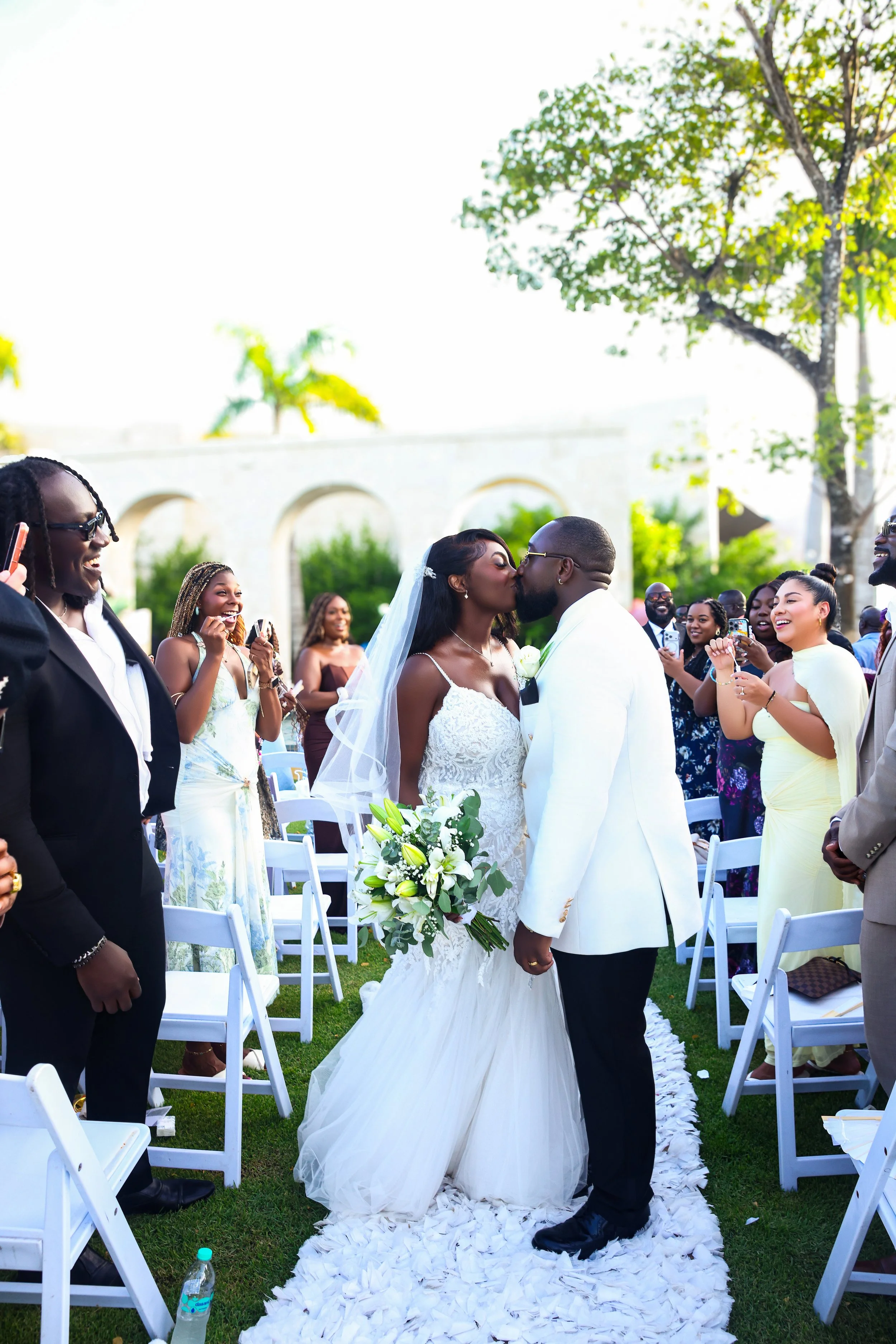 A bride and groom kiss at their outdoor wedding ceremony in a lush green garden, surrounded by cheering guests. The bride is wearing a white lace wedding dress with a veil, holding a bouquet of white flowers, while the groom is in a white tuxedo. Gue