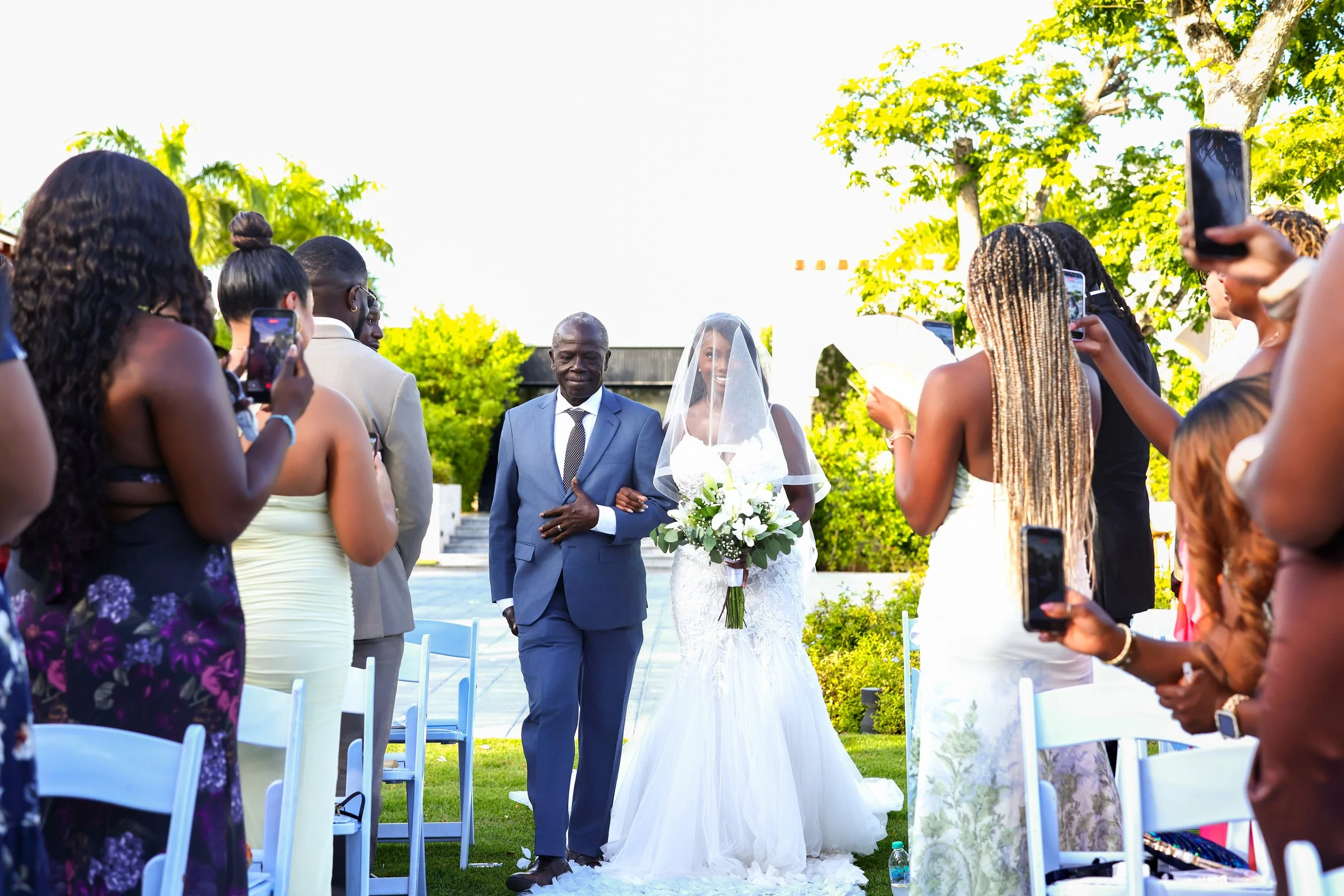 A bride and an older man walking down the aisle at an outdoor wedding, surrounded by guests taking photos. The bride is holding a bouquet and wears a veil; the man wears a gray suit. The setting is lush with green trees, and sunlight filters through 