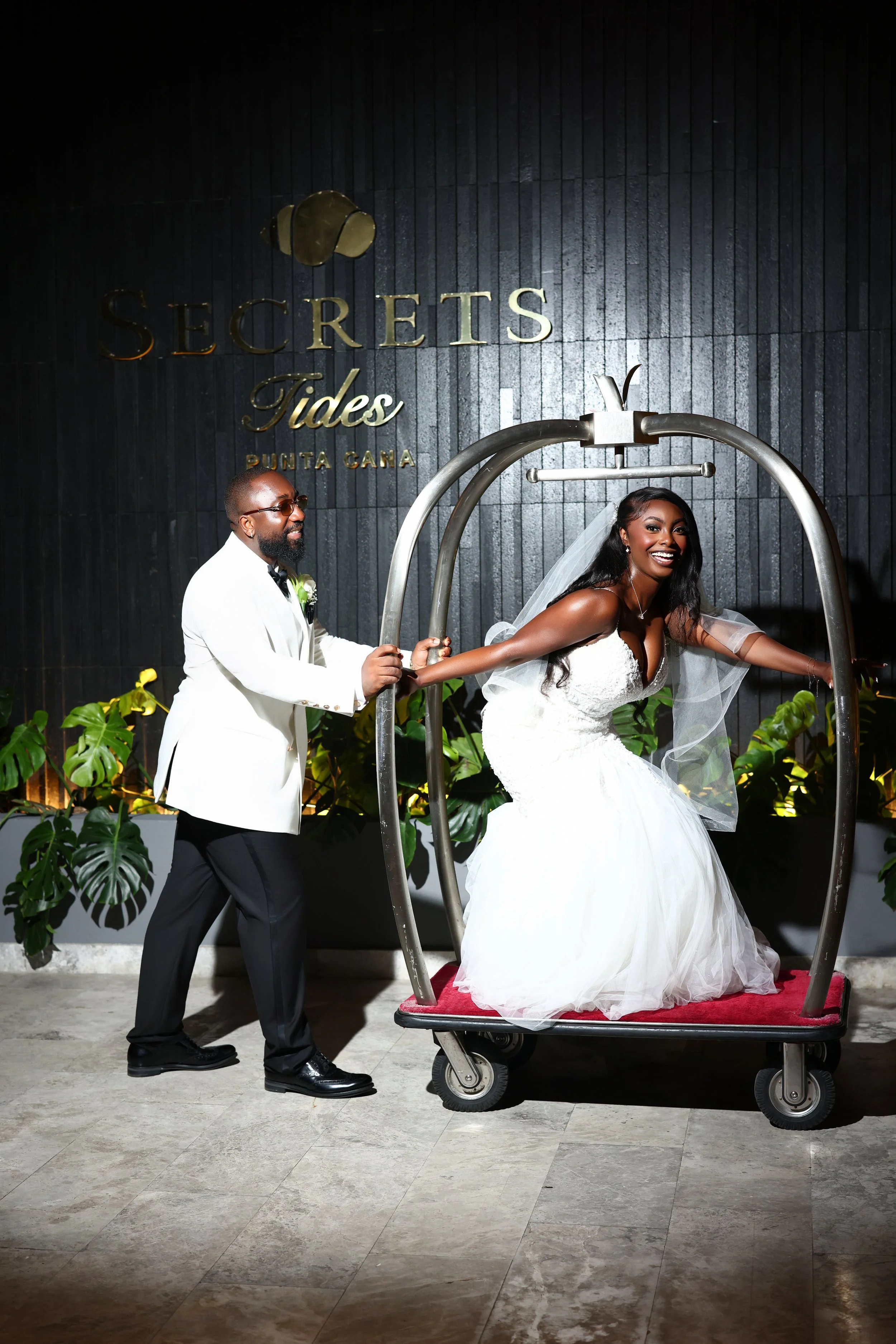 A bride in a white wedding dress and veil sitting on a red carpeted hotel room cart, smiling while holding onto the handle, with a man in a white tuxedo standing beside her, holding her hand. The background features a black wall with gold lettering t