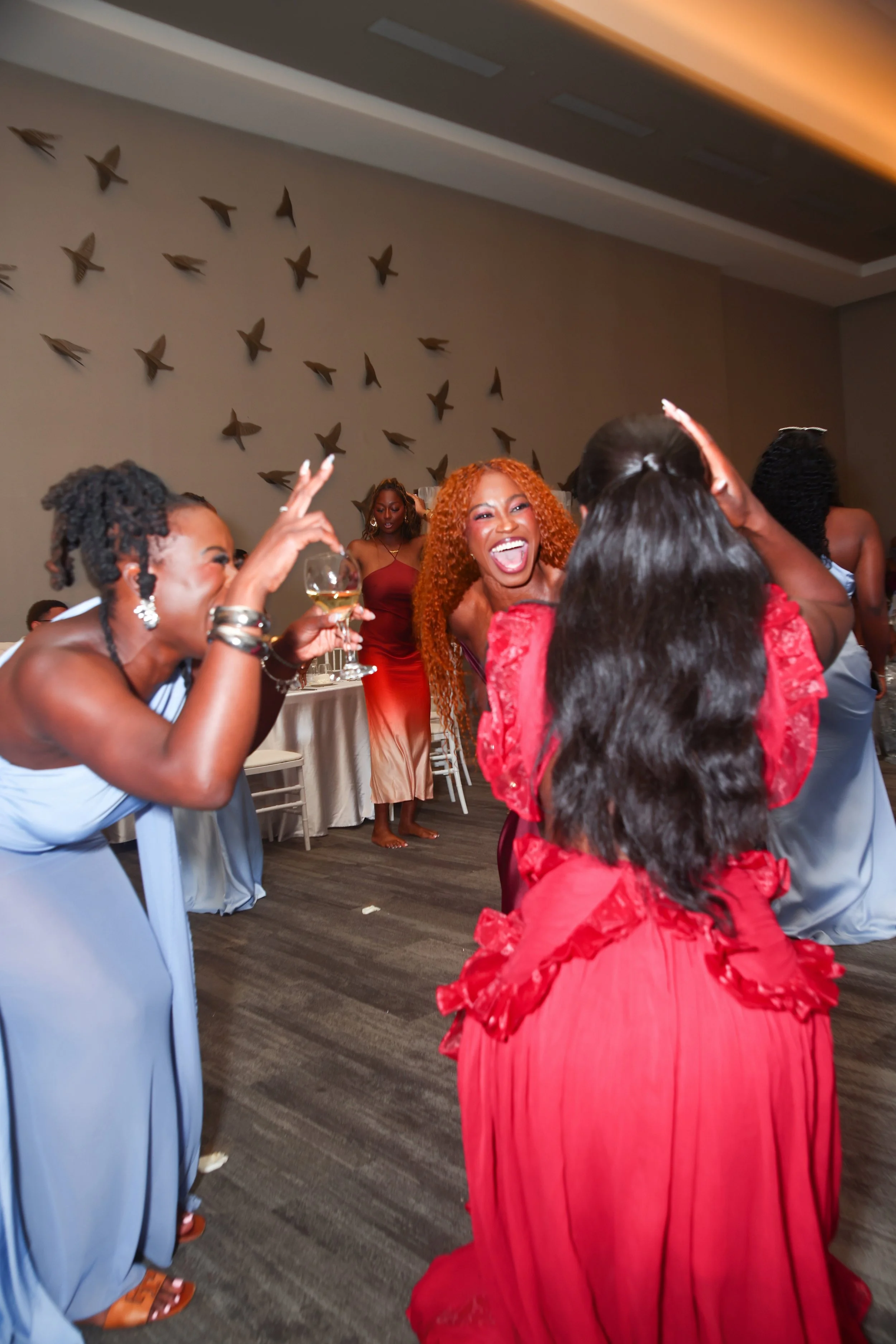 A group of women celebrating at a party or wedding reception. They are dancing, laughing, and enjoying each other's company. Some are holding drinks, and one woman is seen in the background standing near a decorated wall with bird ornaments.
