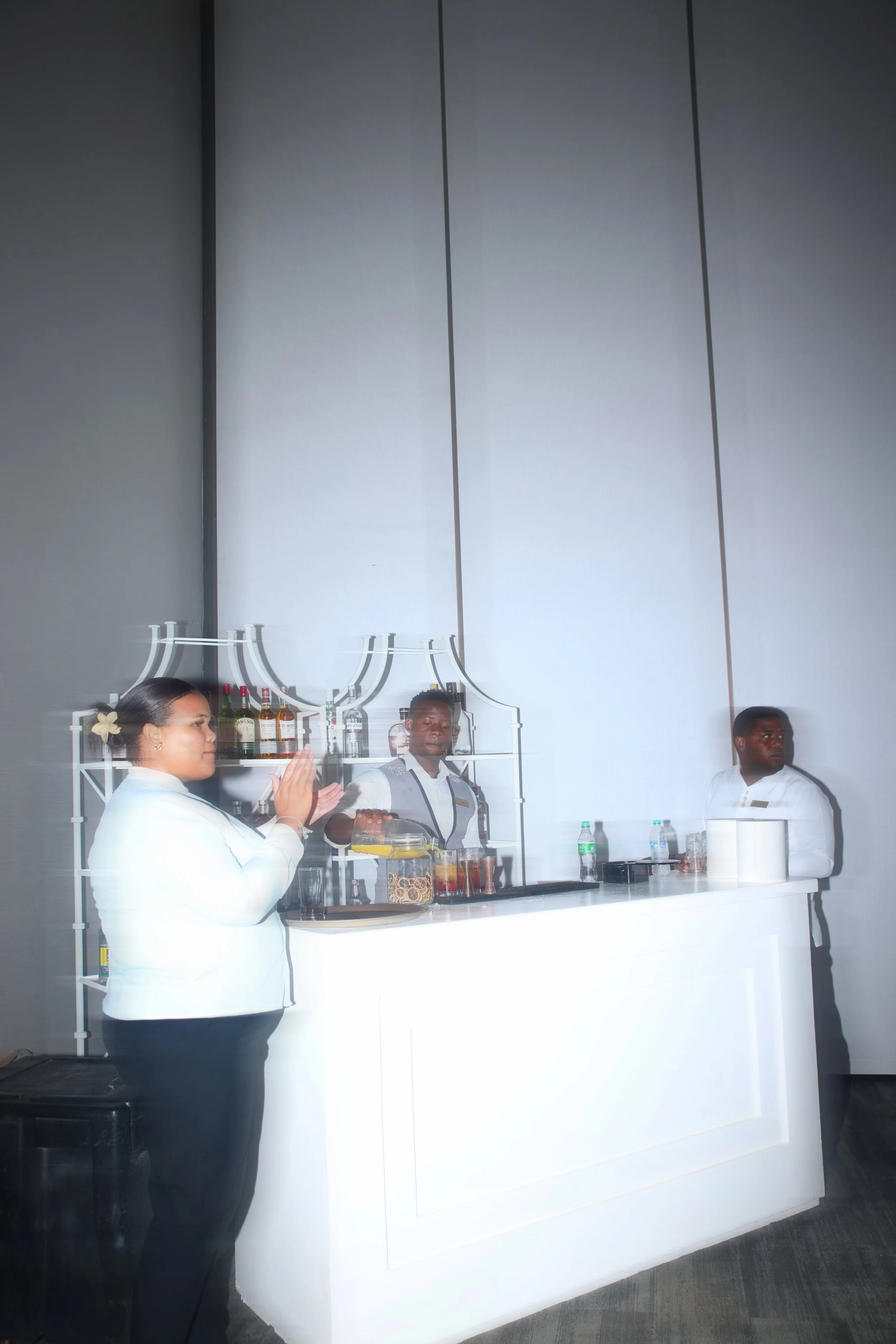 Three bartenders standing behind a white bar, with bottles of alcohol and glasses, in a modern, minimalistic bar or event space with a gray wall and a large white counter.