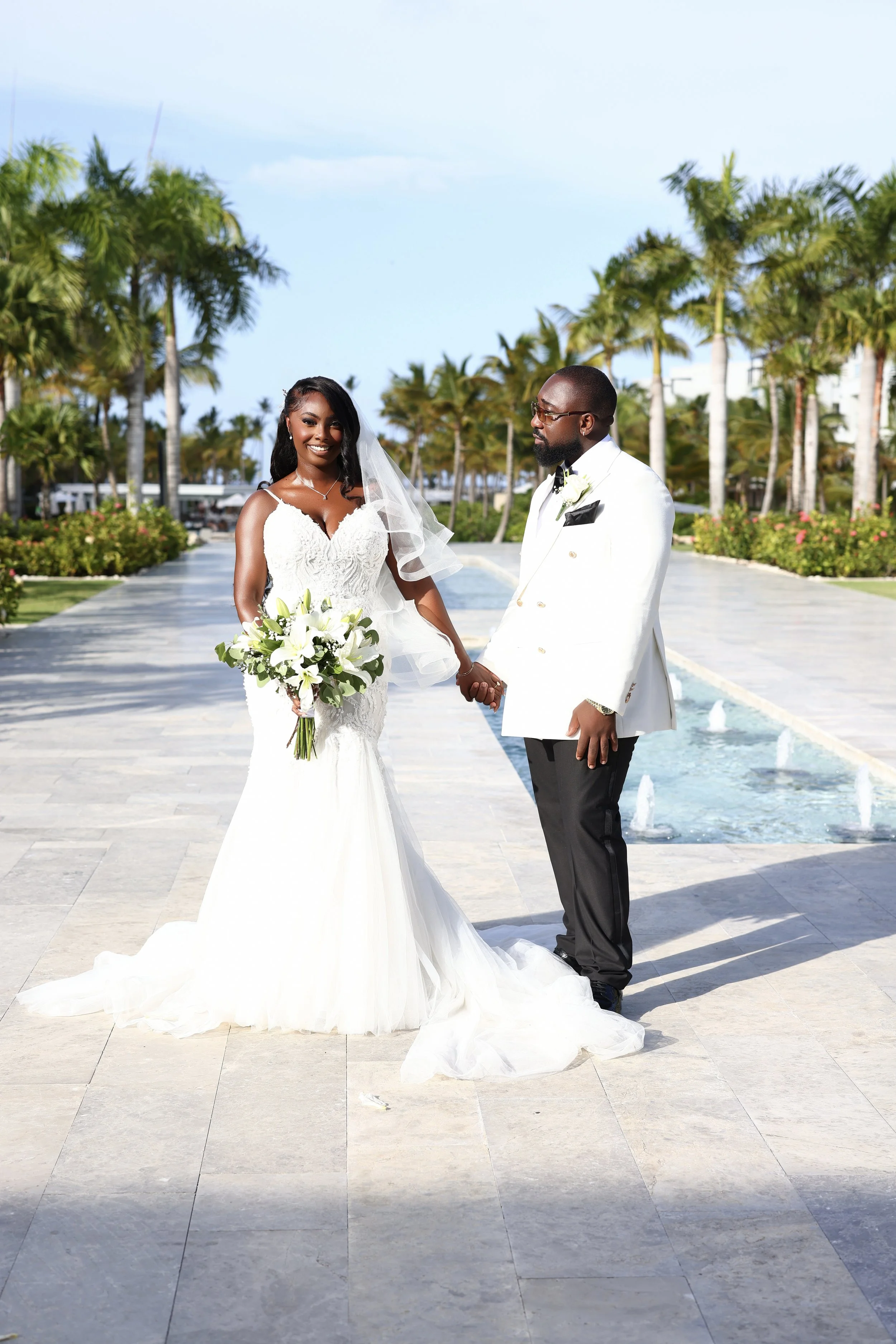 A bride in a white wedding dress holding a bouquet of white flowers stands next to a groom in a white tuxedo jacket with black pants, on a paved outdoor walkway lined with palm trees, with a fountain running along the sides, under a clear blue sky.
