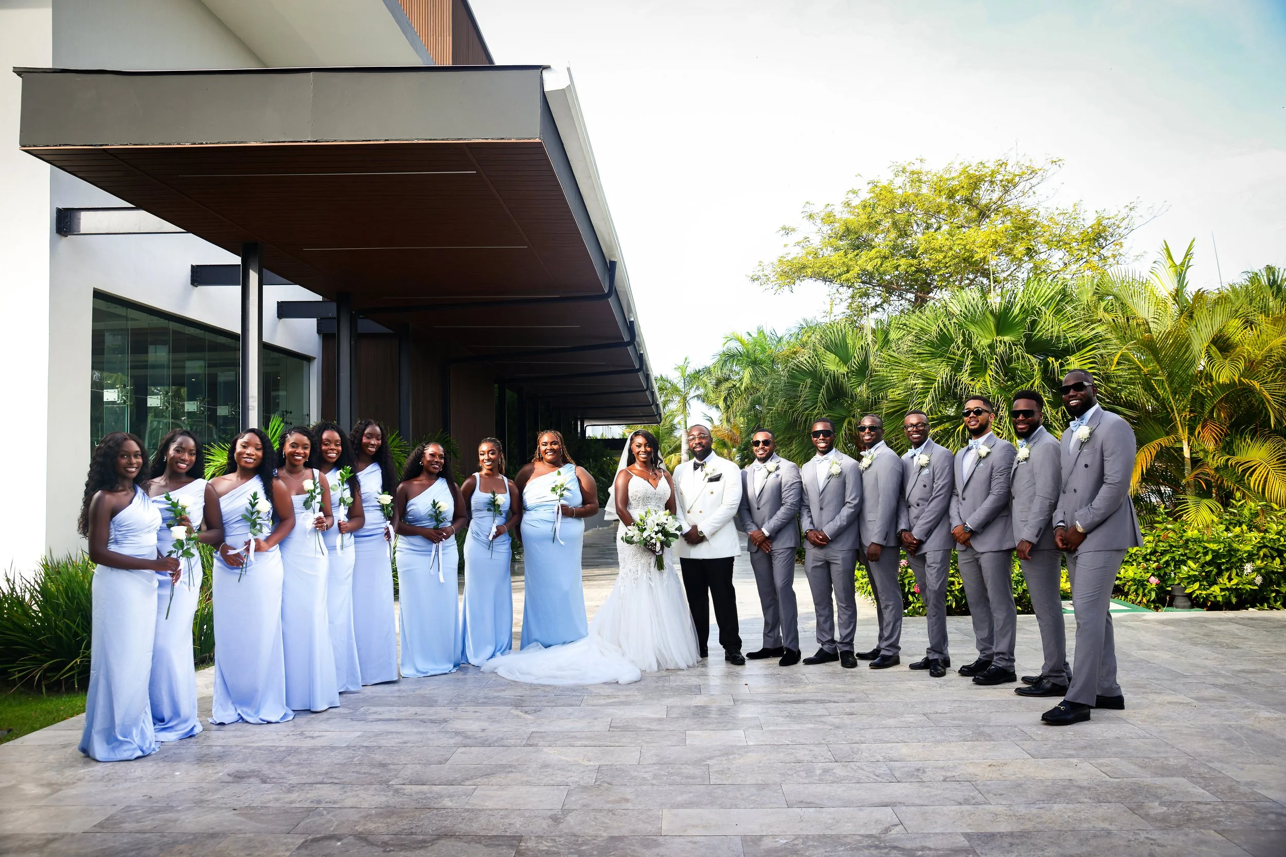 A group of men and women dressed in formal wedding attire standing outdoors near a modern building with trees and greenery in the background.