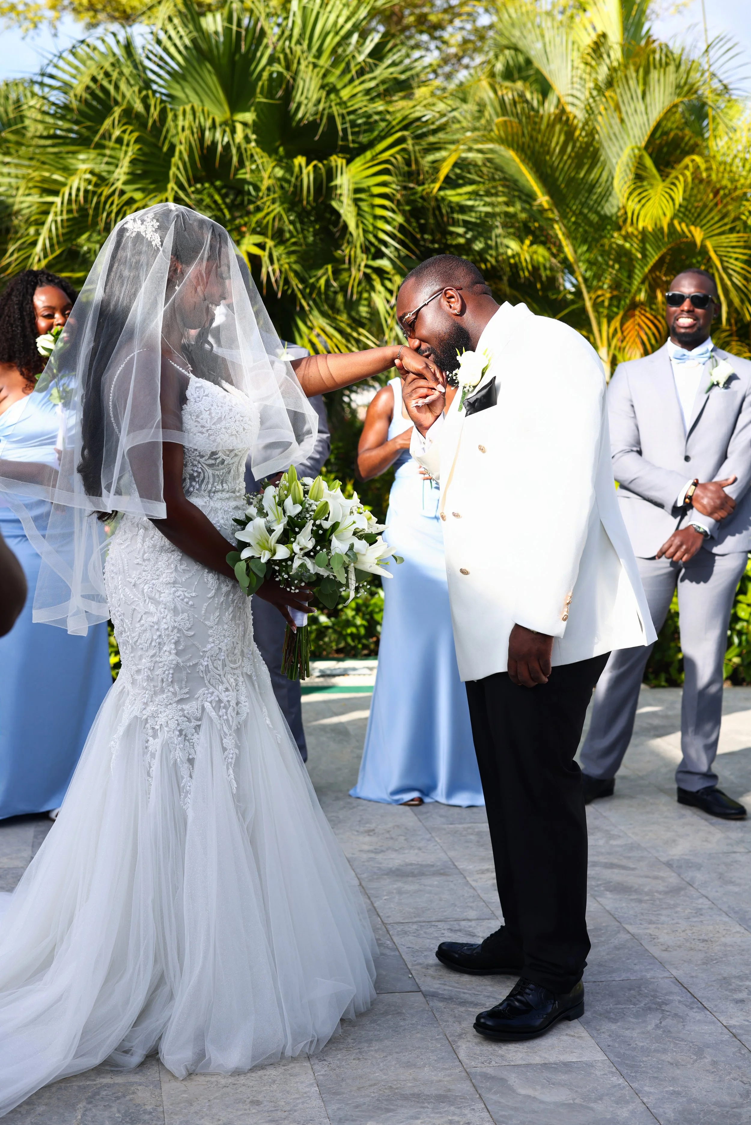 A bride and groom share a kiss during their outdoor wedding ceremony surrounded by friends and family. The bride is wearing a lace wedding gown and veil, holding a bouquet of white lilies. The groom is dressed in a white tuxedo jacket, black trousers