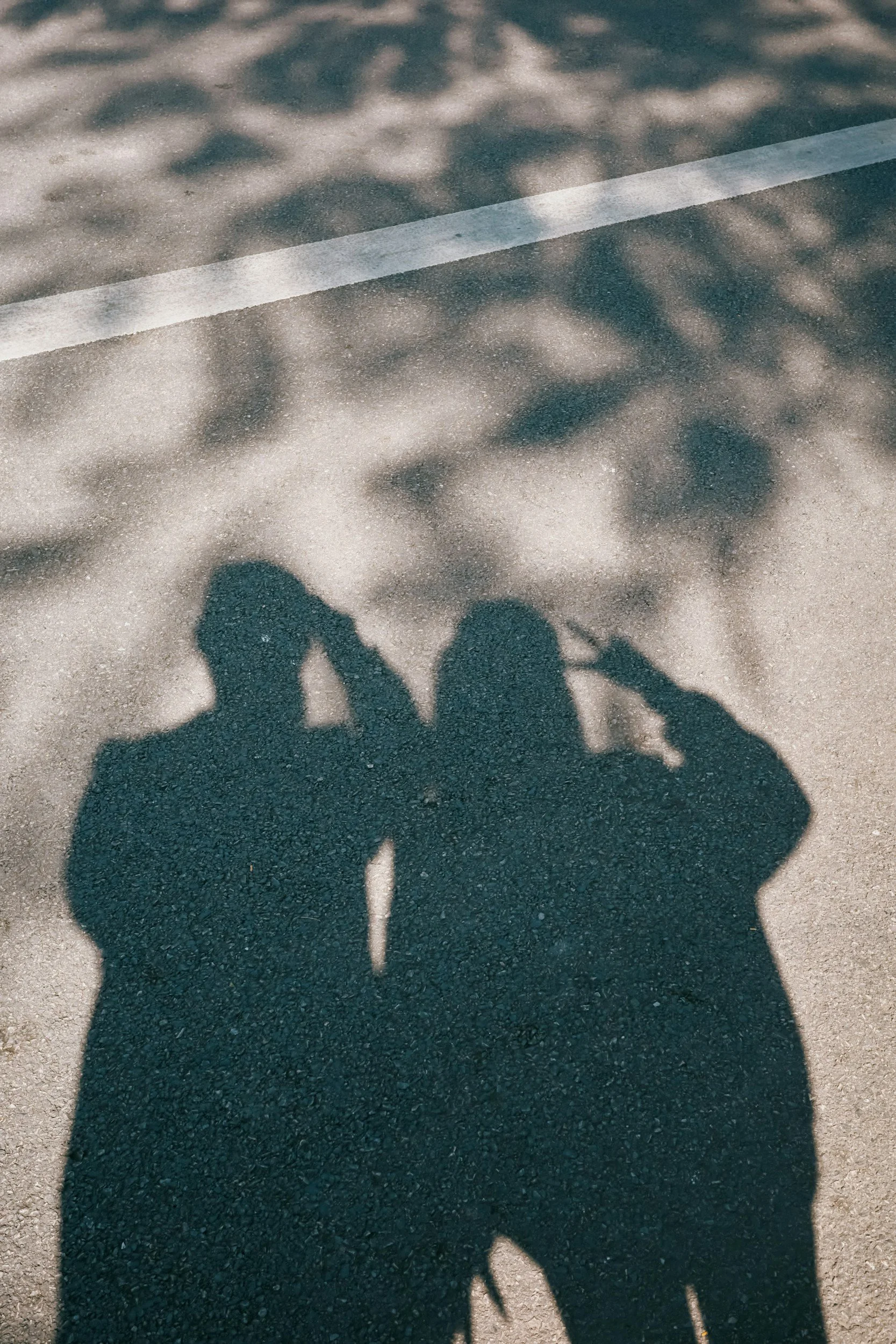 Shadows of two people on the ground, one holding a camera or phone, with dappled sunlight and a white line on the pavement.