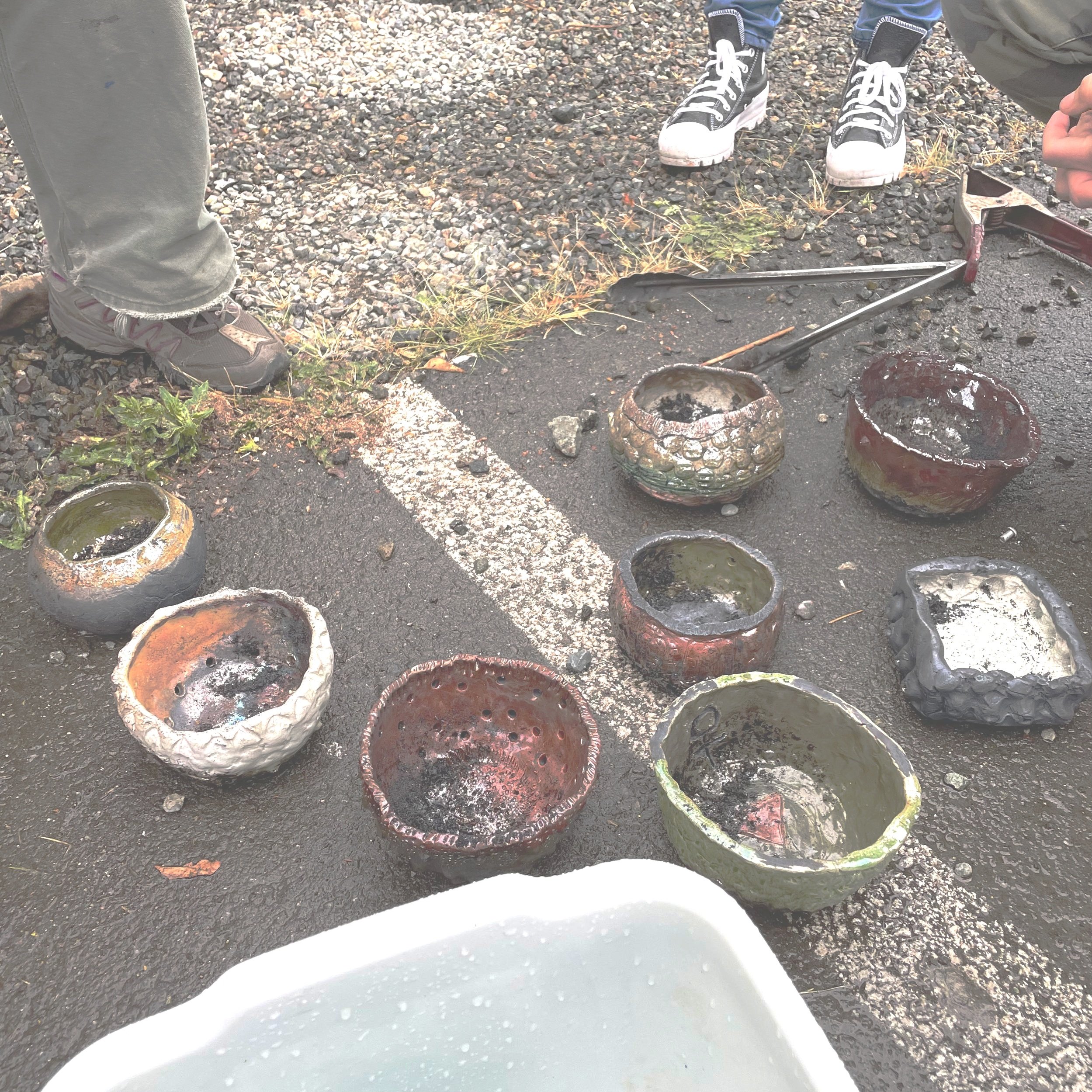 A collection of small, colorful, handcrafted ceramic bowls and containers on an outdoor asphalt surface. Some bowls appear glazed with shiny finishes, and a few have holes or textured surfaces. Two people are standing nearby, one wearing gray pants and the other wearing blue jeans and black high-top shoes. A metal工具 is also present on the ground.