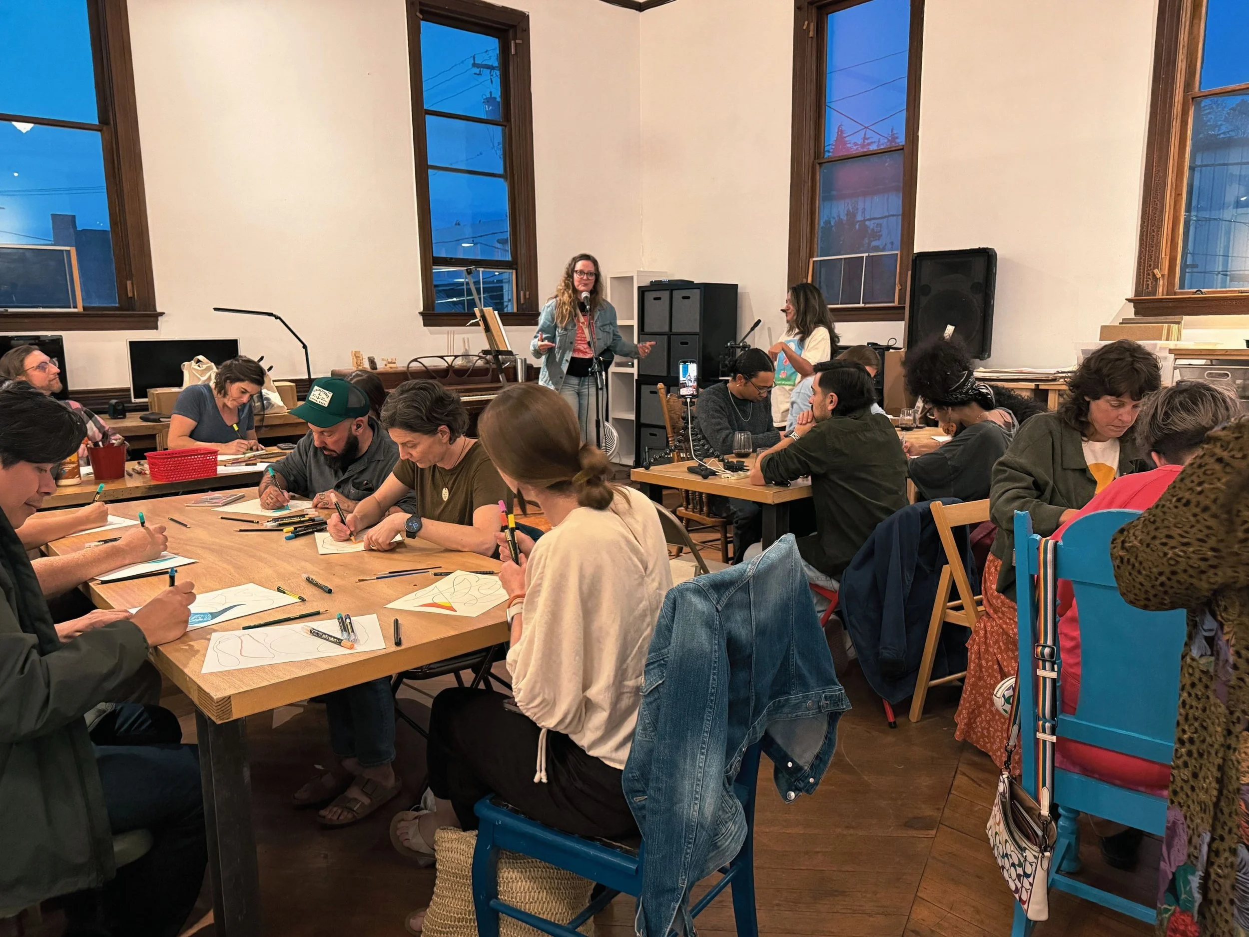 A group of people sitting at tables in a room during a workshop or class, with two women standing at the front, one speaking and the other preparing to speak, surrounded by computer monitors, books, and audio equipment.