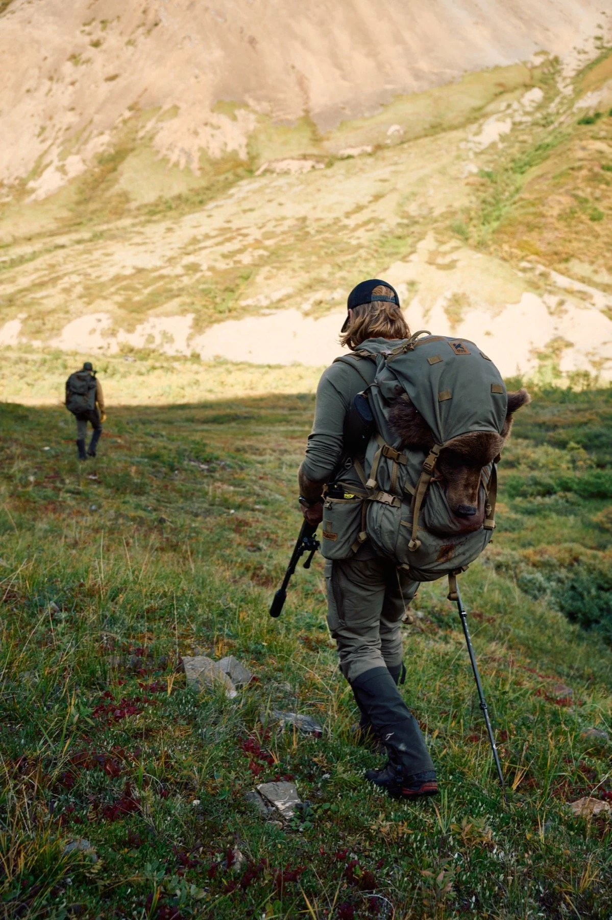 Hunter packing out a grizzly with a rifle and trekking pole down a sunlit valley, his partner ahead