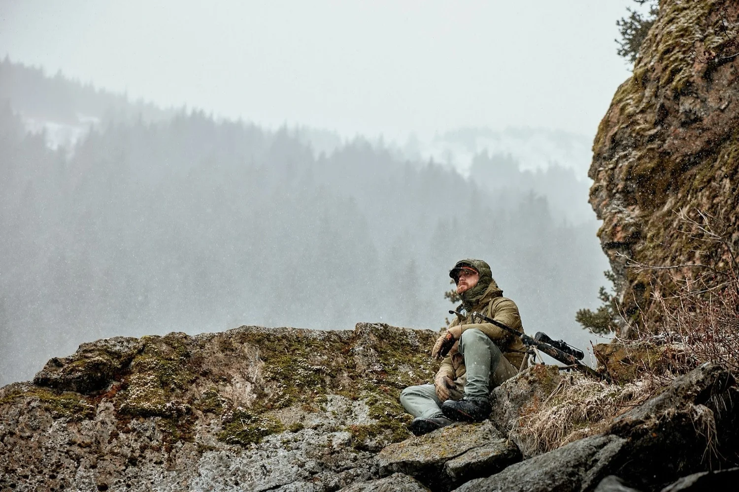 Hunter sitting on a rocky ledge with a rifle above a foggy, snowy valley