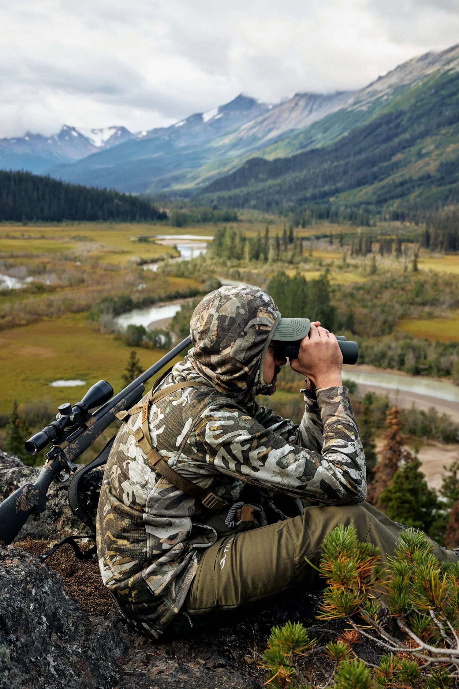Hunter in camo glassing with binoculars from a rock, rifle beside him, river valley and peaks behind