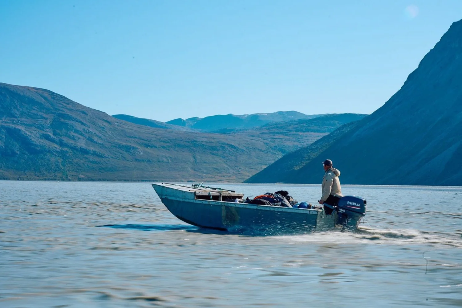 Small open boat running across a fjord below mountains, one person at the helm