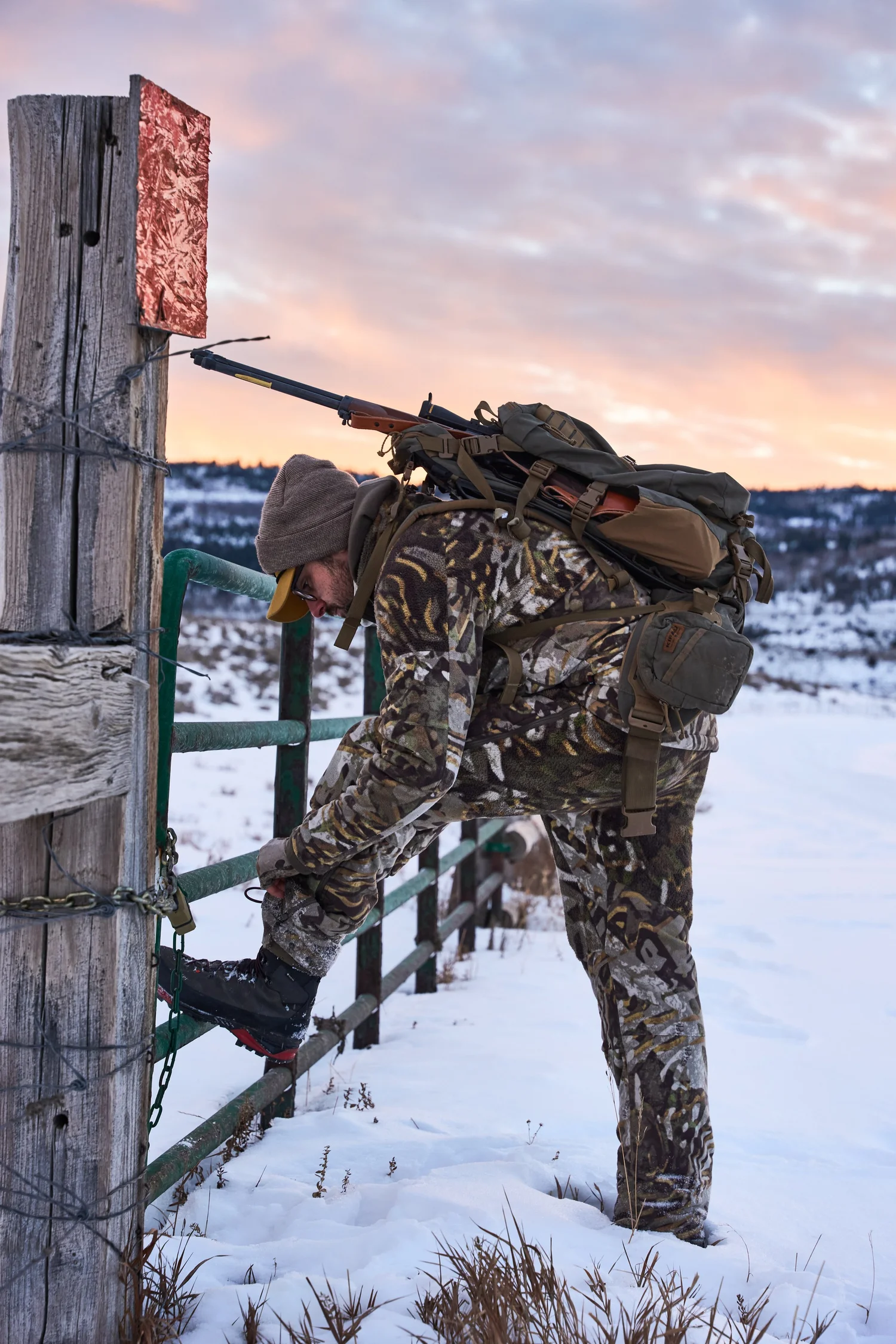 Hunter climbing a ranch gate in the snow with a lever-action rifle on his pack, sunrise sky behind