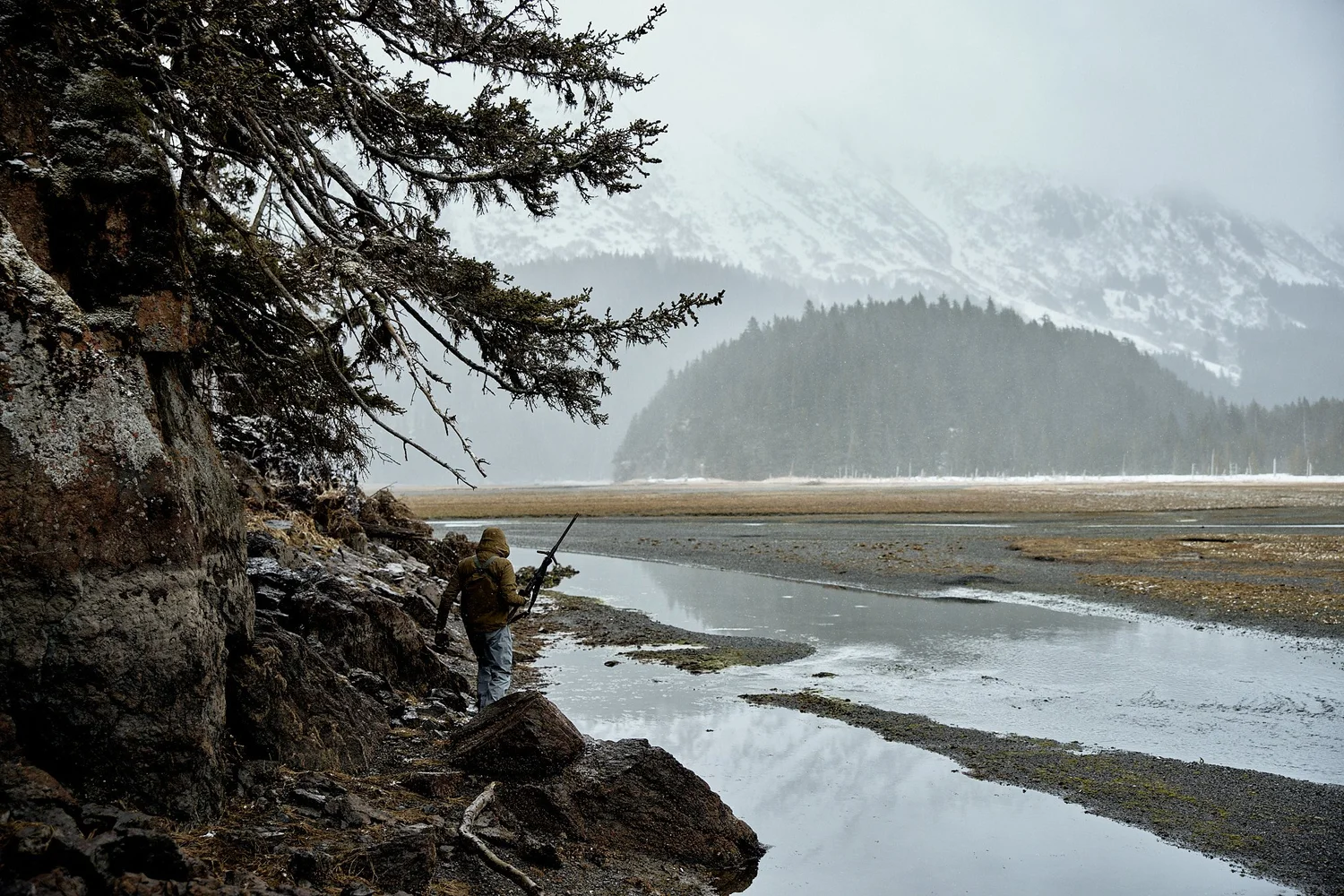 Hooded hunter carrying a rifle along a rocky shoreline at low tide, snowy mountains in fog behind