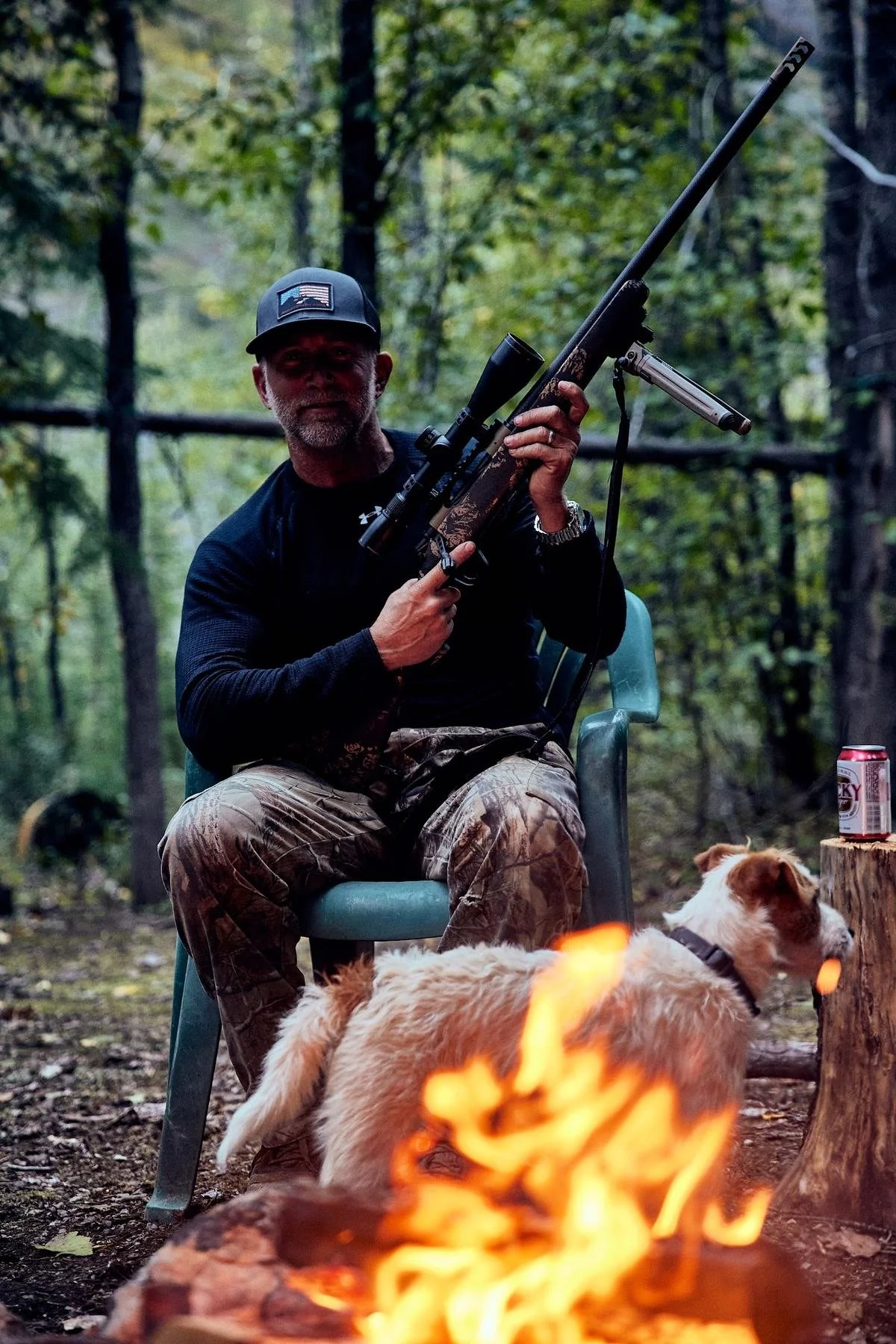 Man in a camp chair holding a rifle beside the campfire, a terrier at his feet