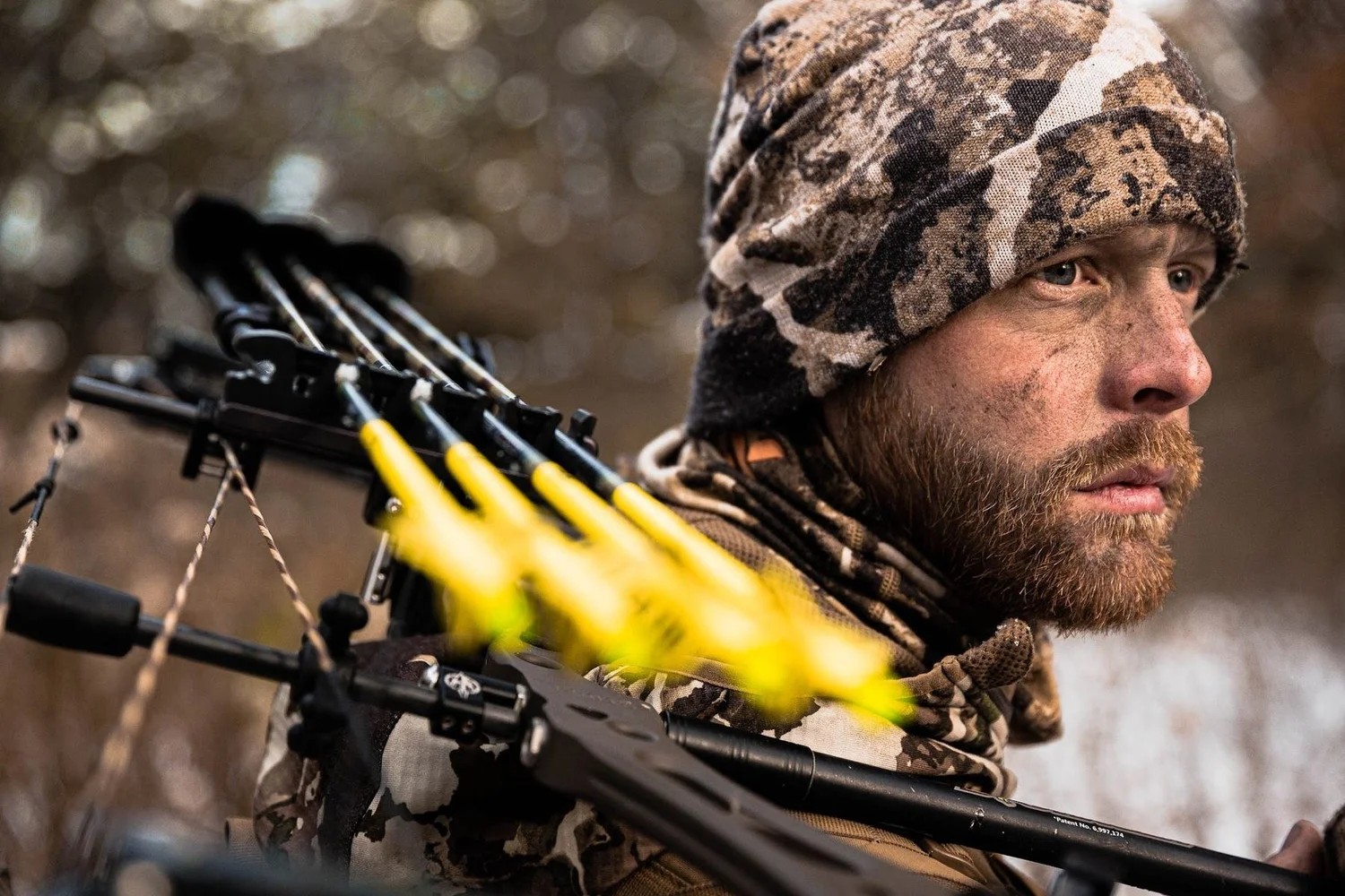 Bearded bowhunter in a camo beanie with a compound bow and yellow-fletched arrows