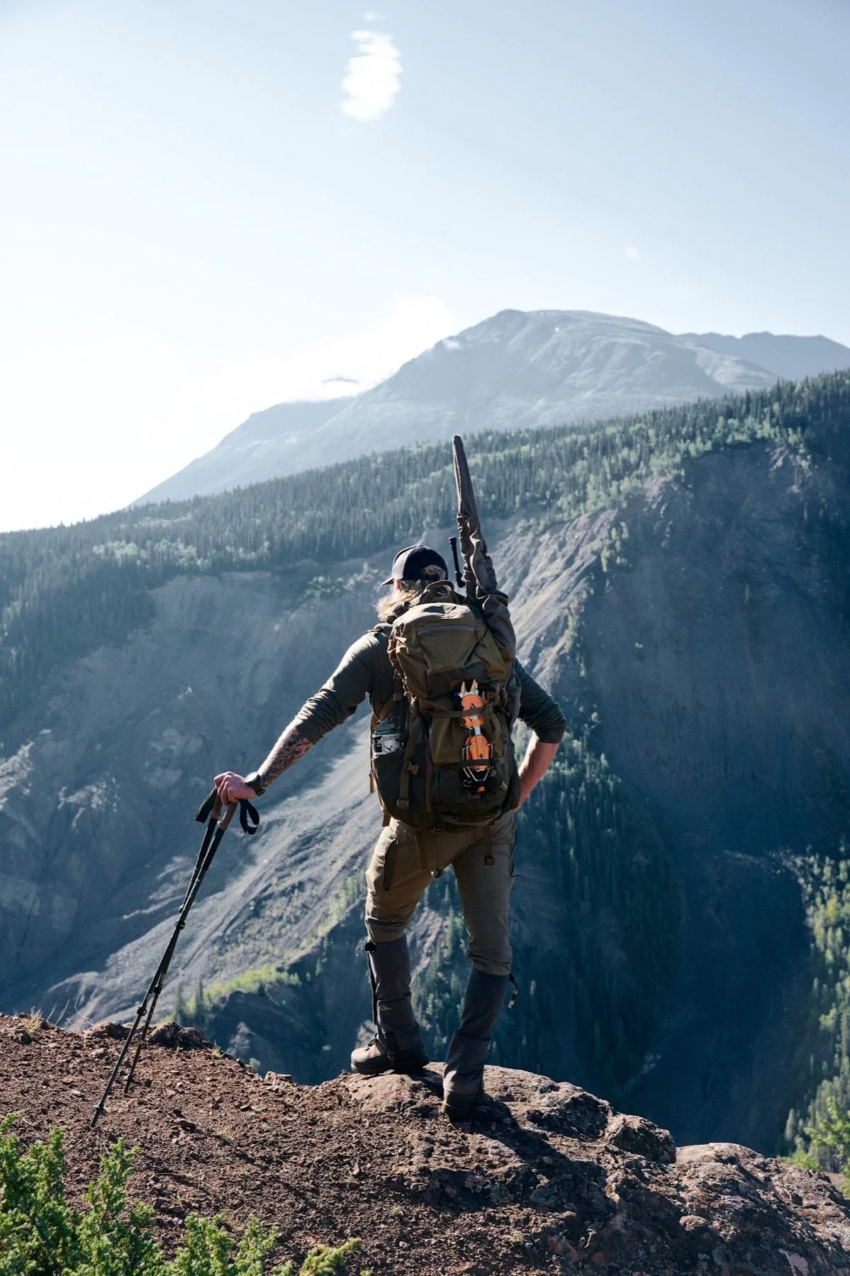 Hunter with a rifle on his pack and a trekking pole on a cliff edge above a canyon
