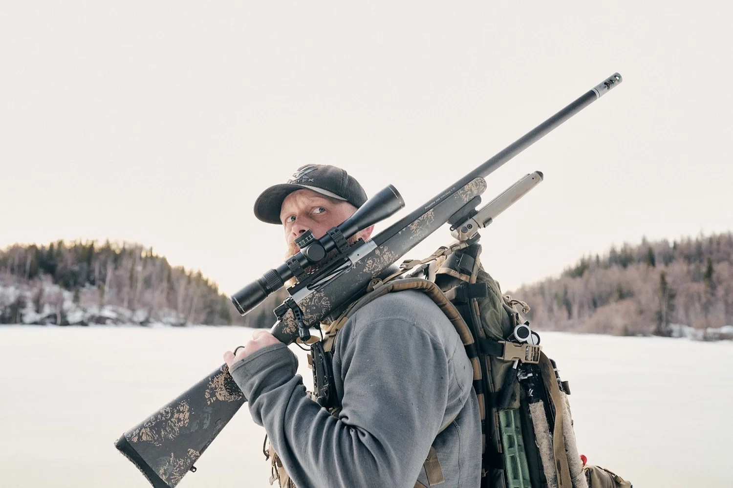 Hunter carrying a scoped rifle on his shoulder, frozen water and white sky behind
