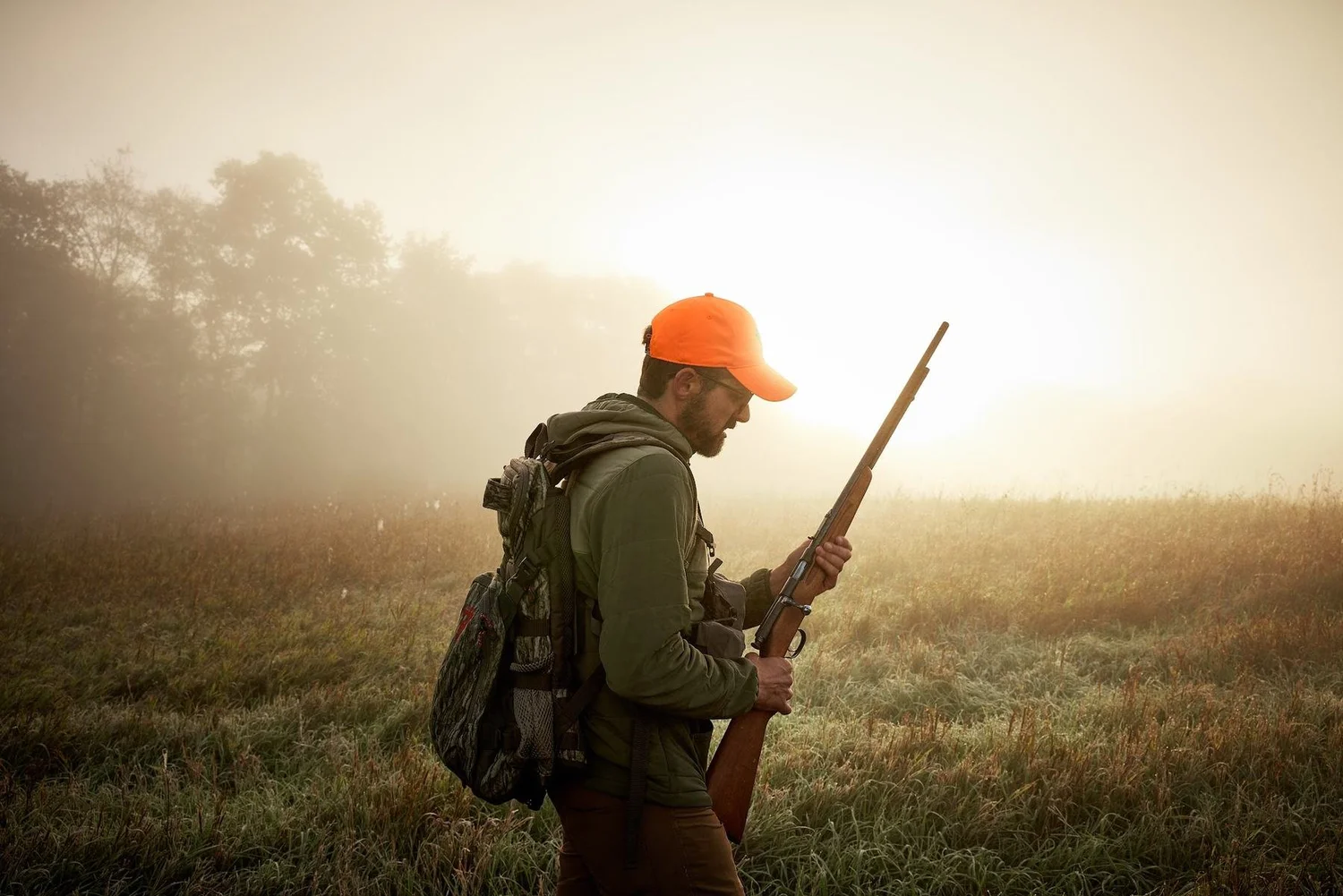 Hunter in an orange cap holding a rifle in a foggy meadow at sunrise