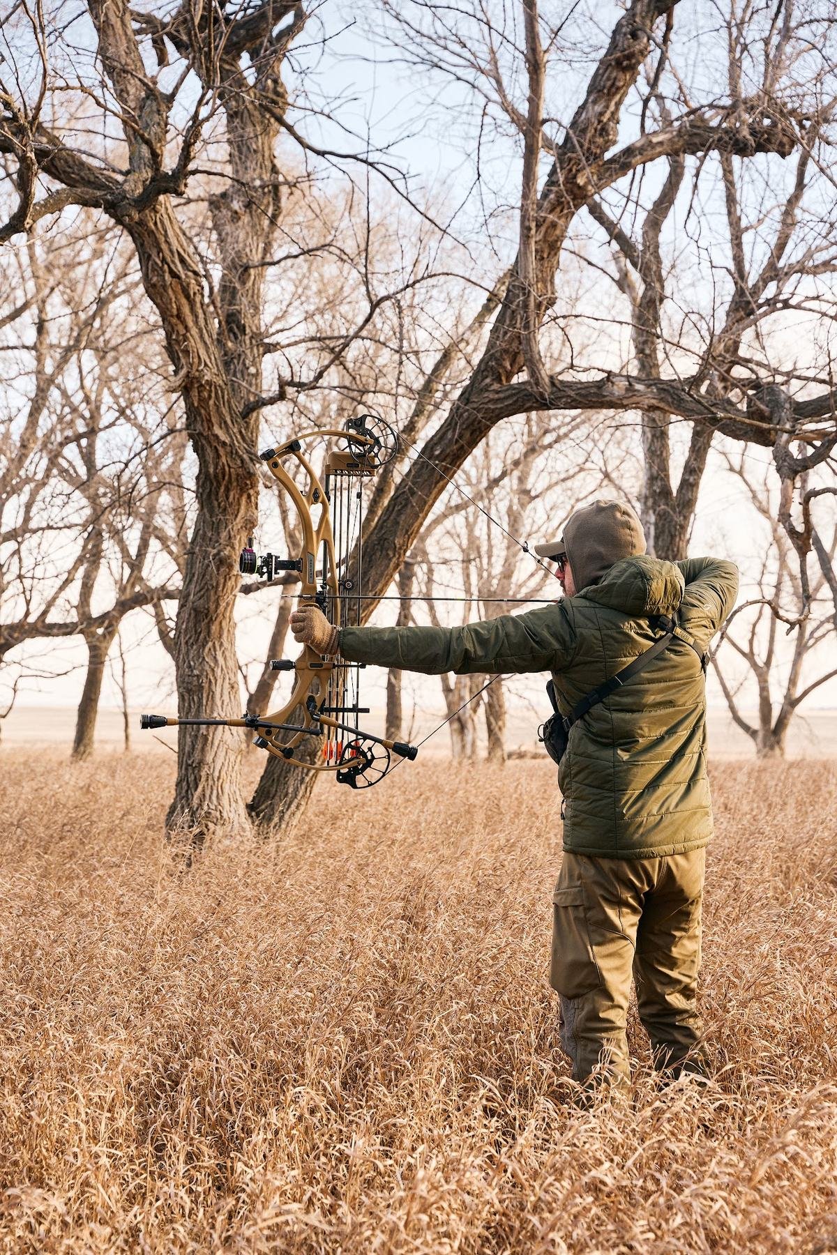 Bowhunter at full draw in tall golden grass under bare trees