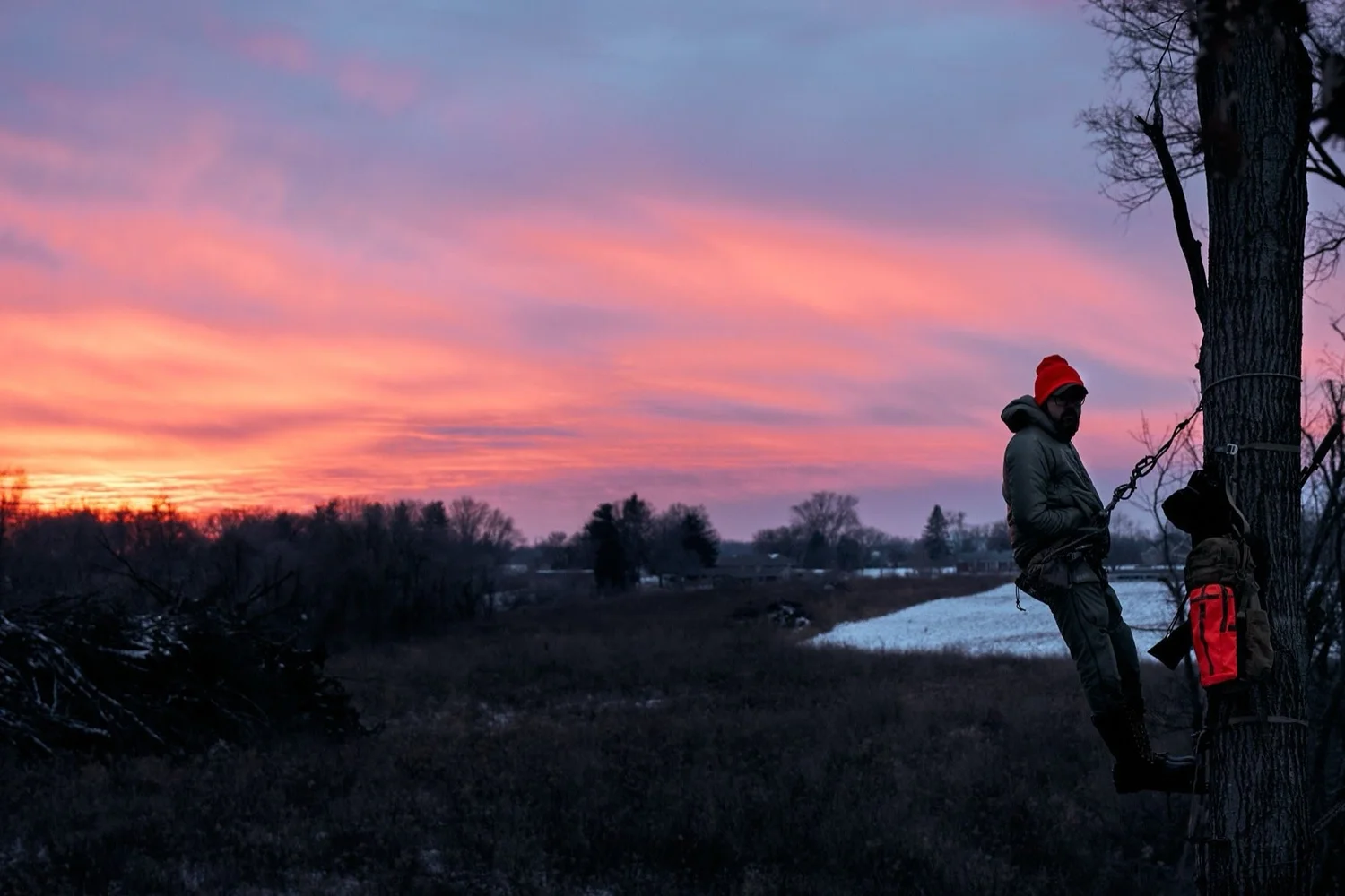 Hunter in a tree saddle against a pink sunset over a snowy marsh