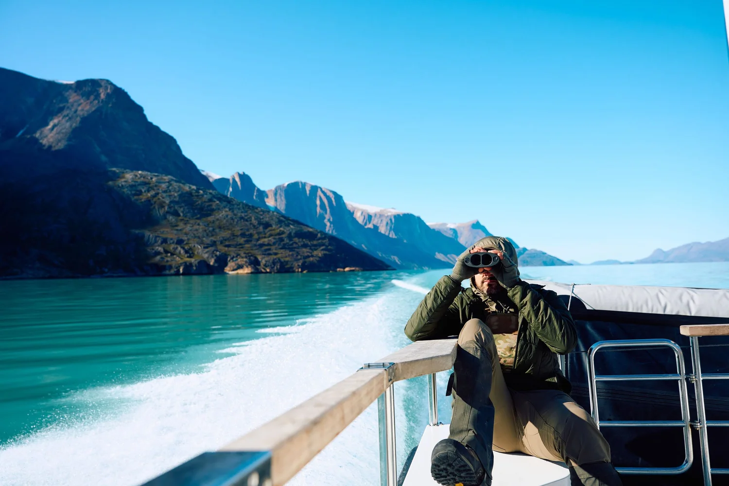 Hunter glassing with binoculars from the boat stern, turquoise fjord and cliffs behind