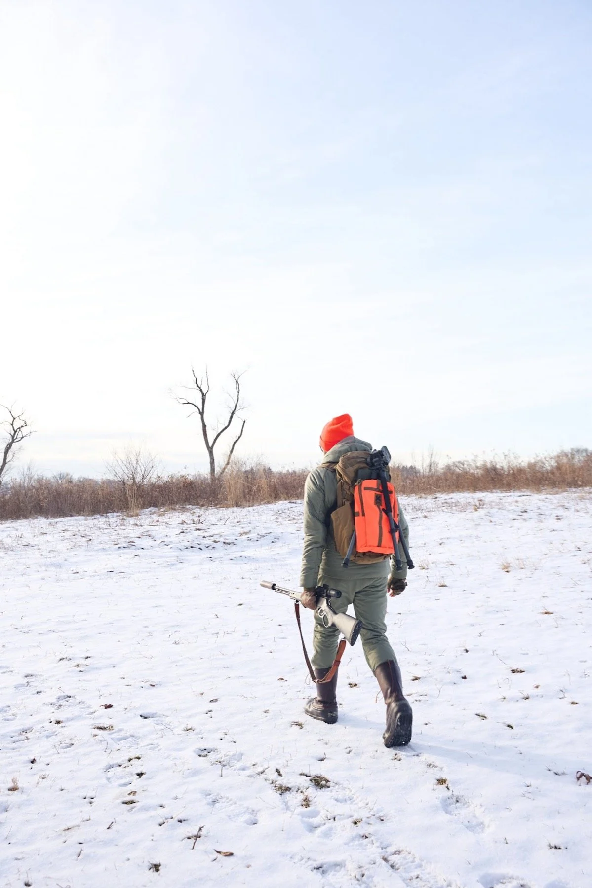 Hunter with an orange pack walking across a snowy field under a bright winter sky