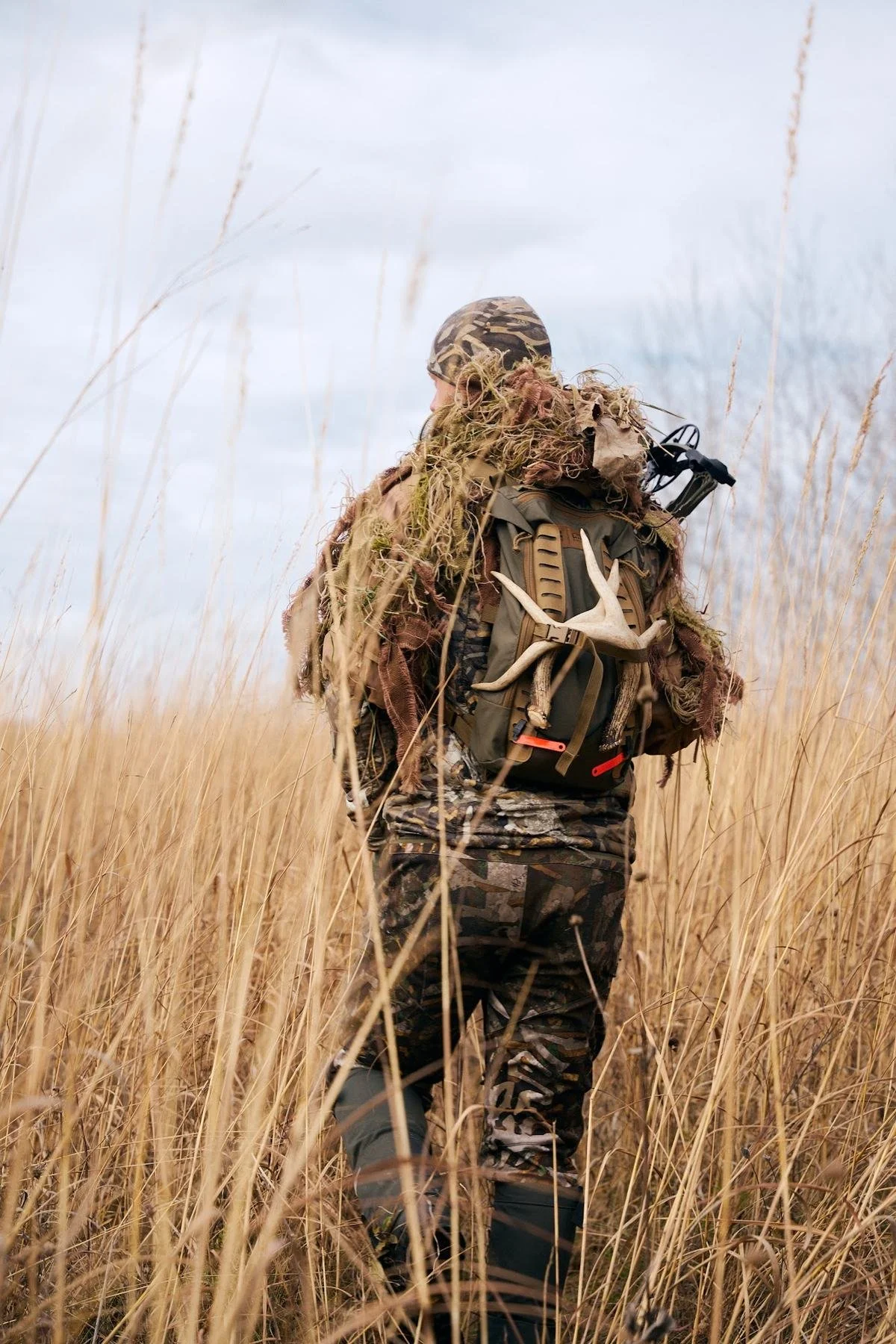 Hunter seen from behind with rattling antlers on his pack, walking through tall golden grass