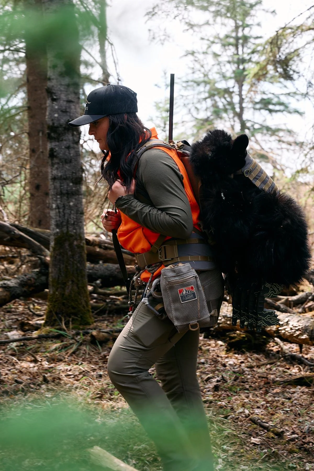 Hunter in a blaze orange vest glancing back at the black bear on her pack in the forest