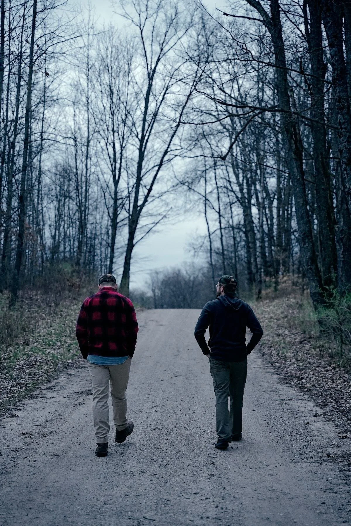 Two hunters walking a gravel road through bare spring woods, seen from behind
