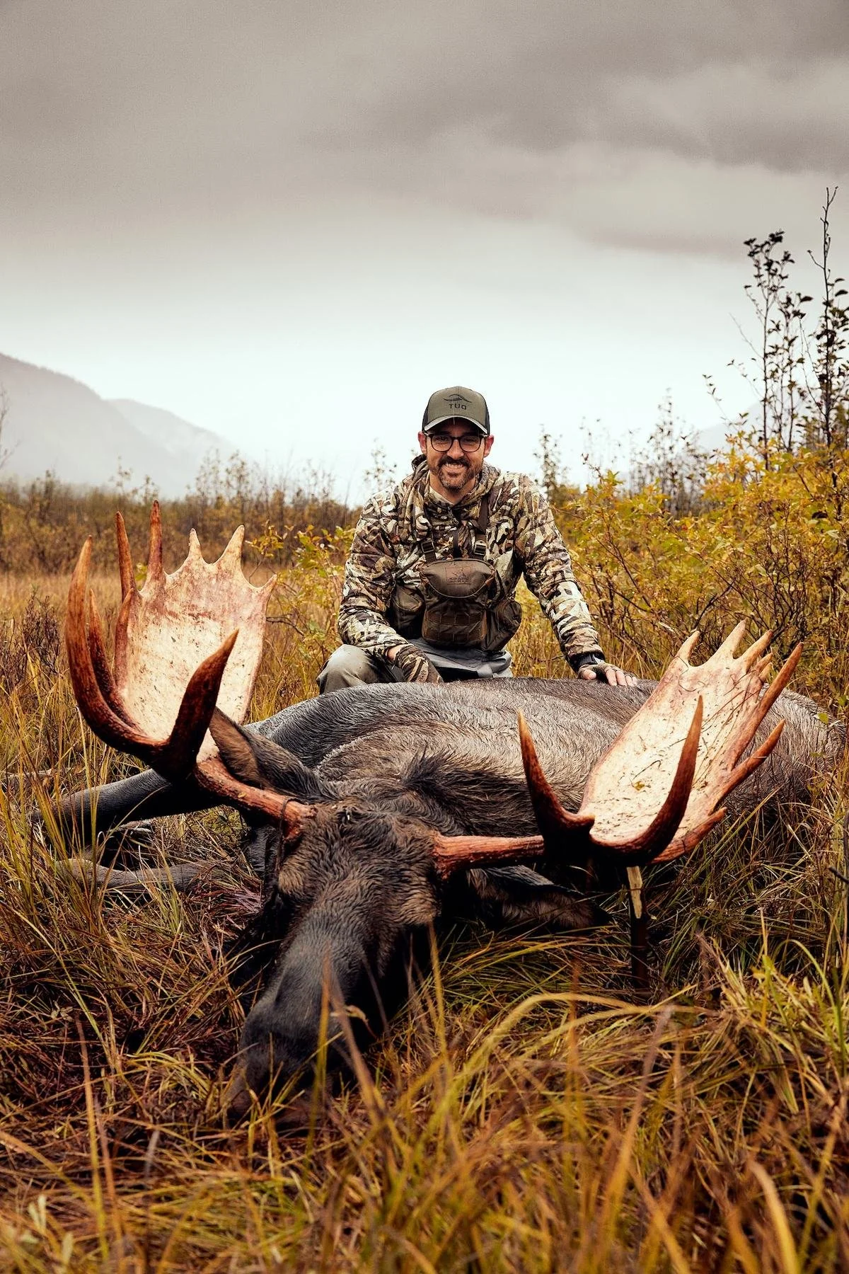 Hunter kneeling behind a bull moose with wide antlers in golden meadow grass