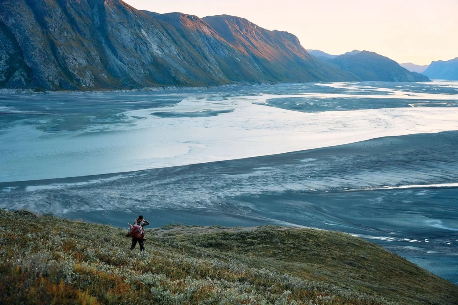 Lone hunter on the tundra above a braided glacial river, mountain cliffs lit pink