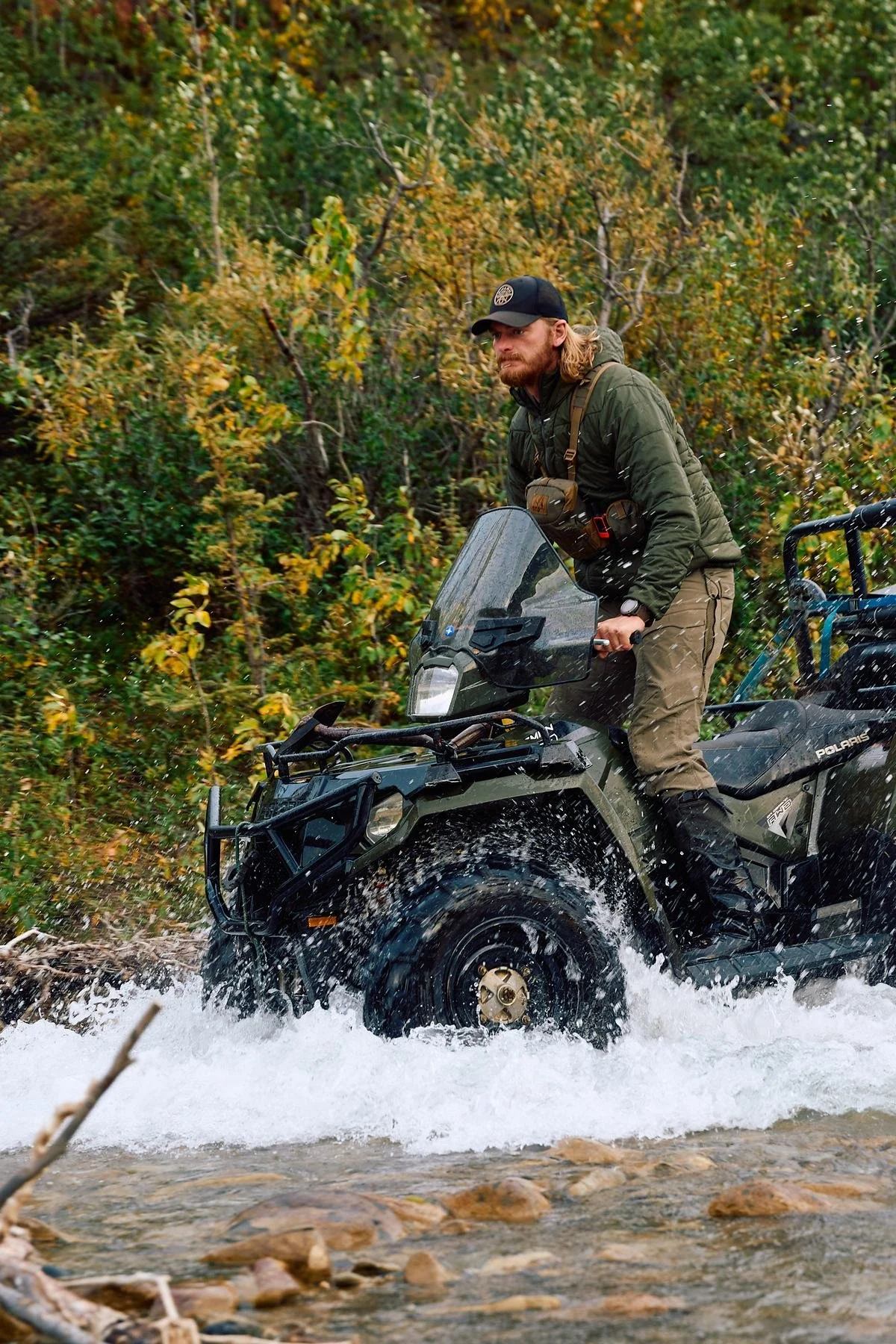 Hunter standing on an ATV as it crashes through a rocky creek, autumn birch behind
