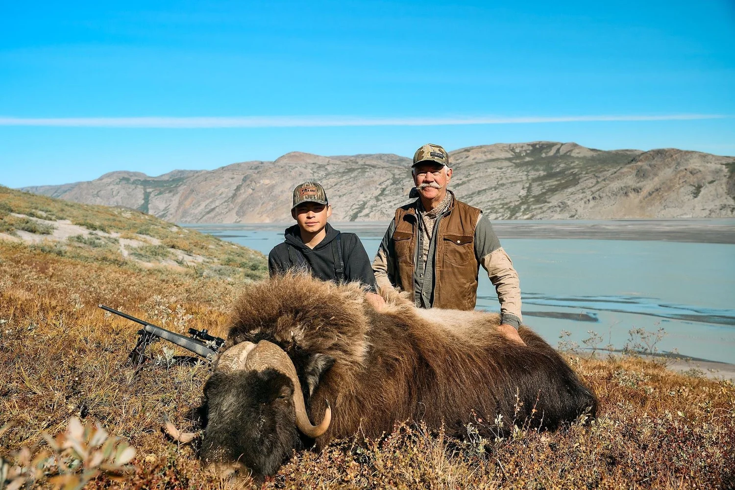 Two hunters kneeling behind a muskox on the tundra, a rifle beside them and the fjord behind