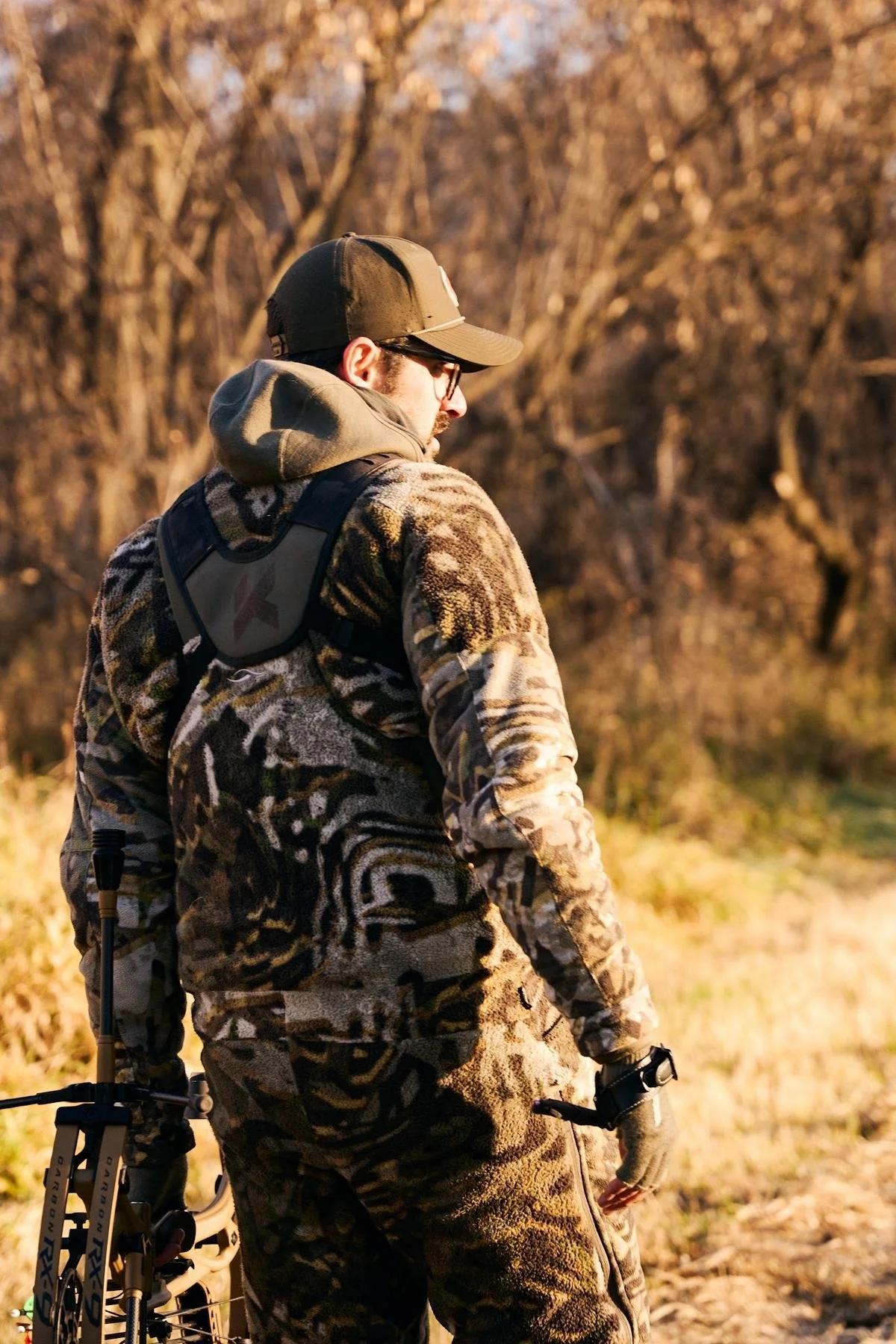 Bowhunter in camo looking back over his shoulder in golden grass at the edge of the woods