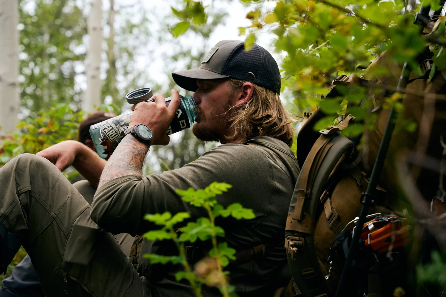 Hunter in a cap drinking from a water bottle while resting in an aspen thicket, a second hunter behind him