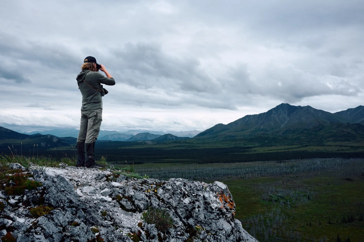 Hunter on a rock outcrop glassing over a wide valley and mountains under heavy cloud