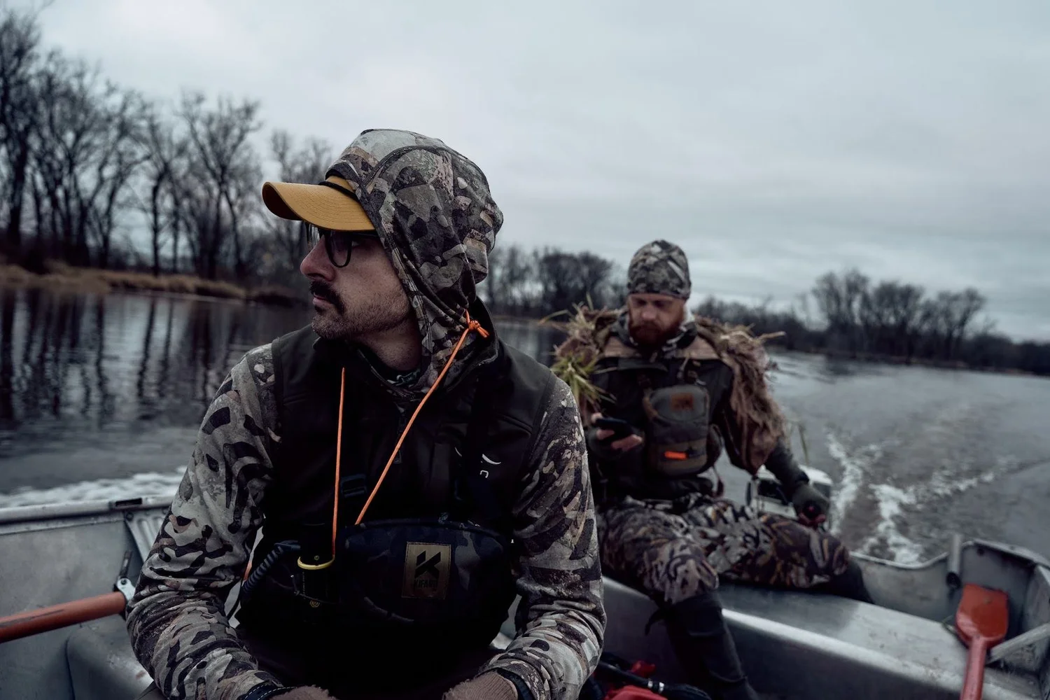 Two hunters riding a jon boat up a river past bare trees