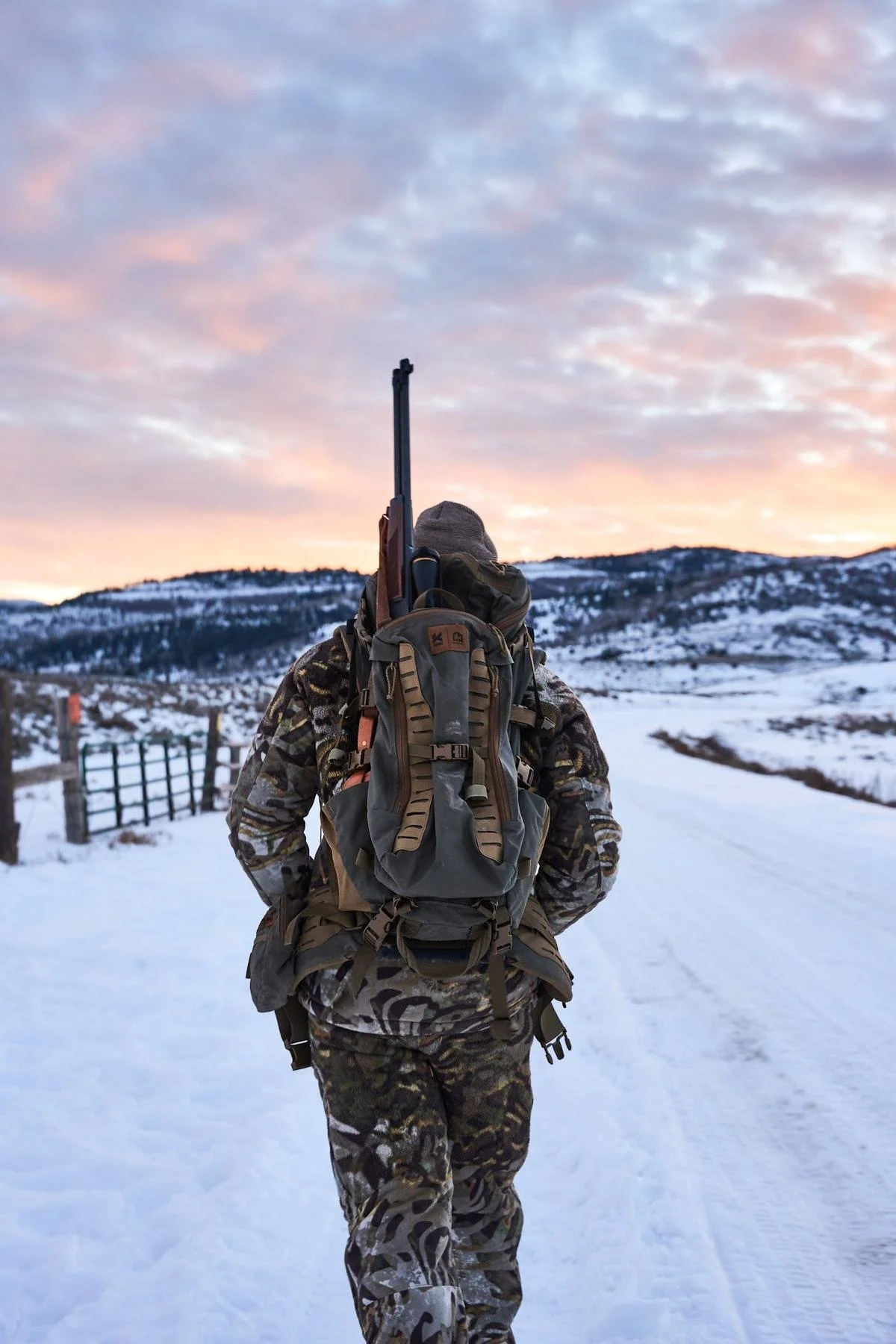 Hunter with a rifle and pack walking a snowy ranch road under pink sunrise clouds