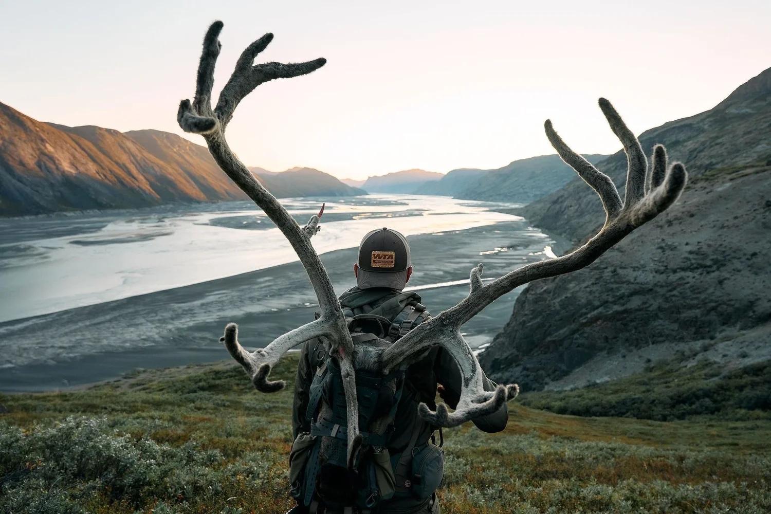 Caribou antlers strapped to a hunter's pack, a braided glacial fjord and sunlit peaks behind