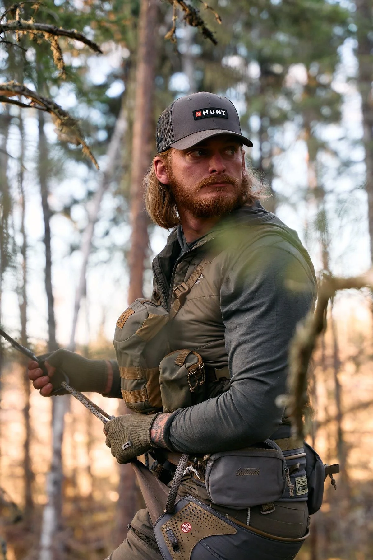 Bearded hunter in a cap pulling up a haul rope at his tree stand, warm light through the pines