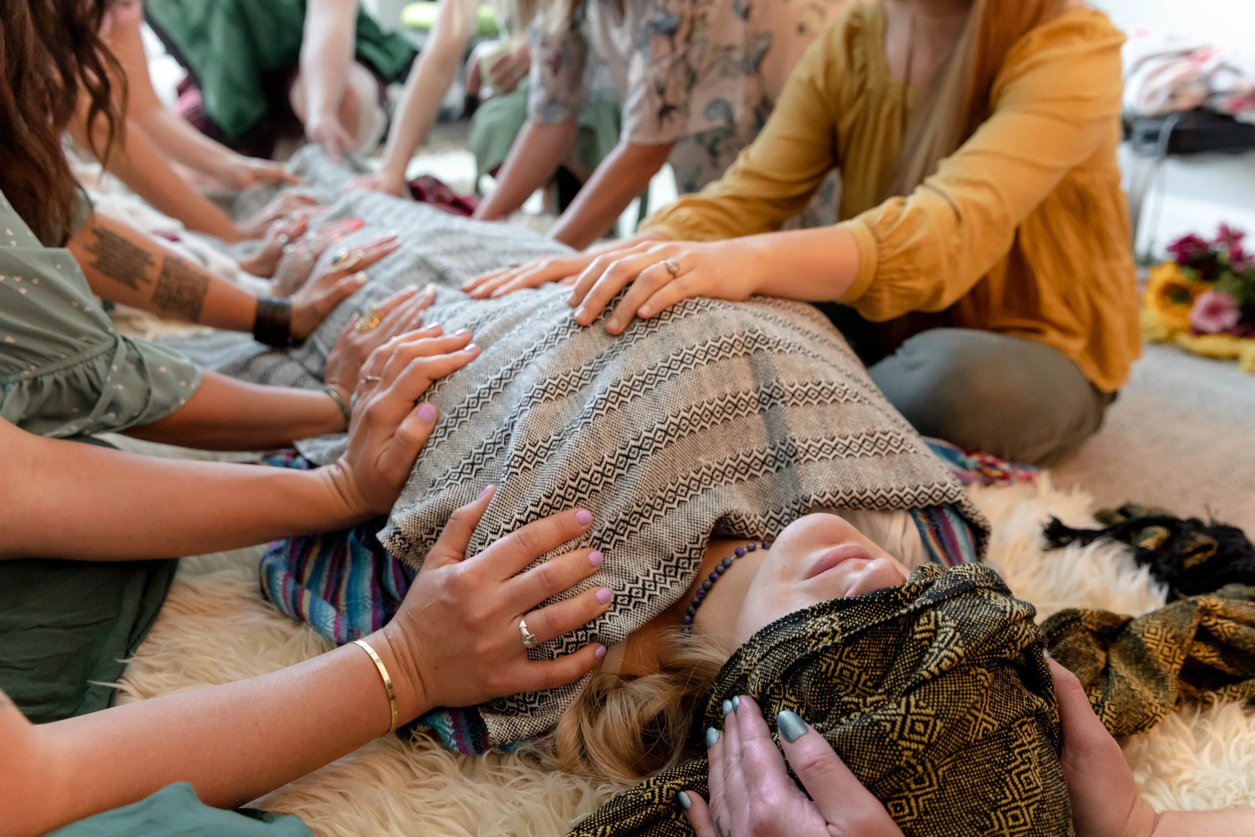 A group of people participating in a Closing of the Bones ceremony around a woman lying on the floor, with hands on her chest and head, in a peaceful setting.
