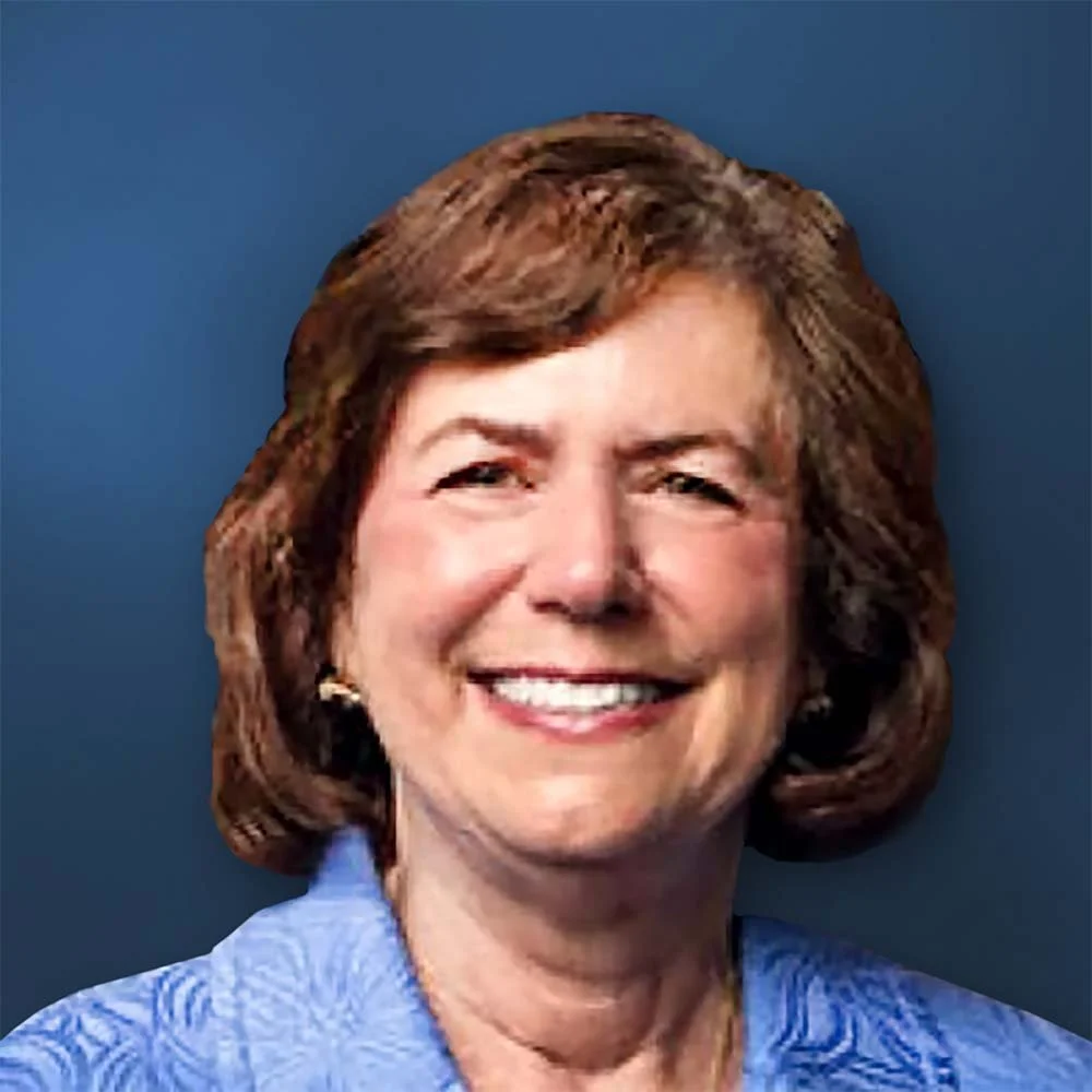 A woman with shoulder-length brown hair smiling at the camera, wearing a blue blazer, in an indoor setting with wooden decor.