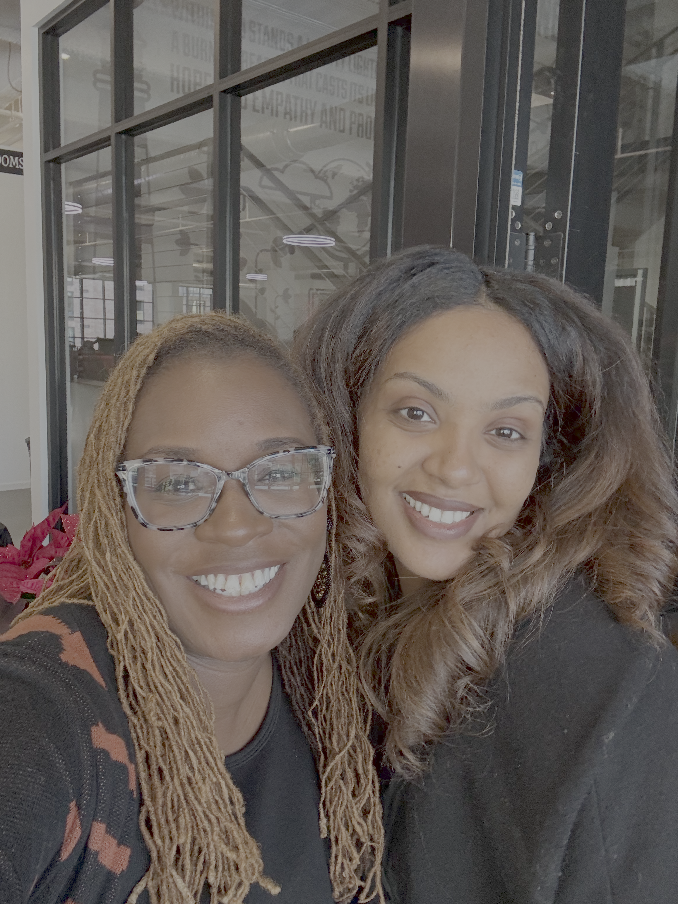 E. Danielle Butler, wearing glasses and locs, and Lulete Mola, smiling together in a selfie inside a modern workspace with inspirational text visible on the glass wall behind them.