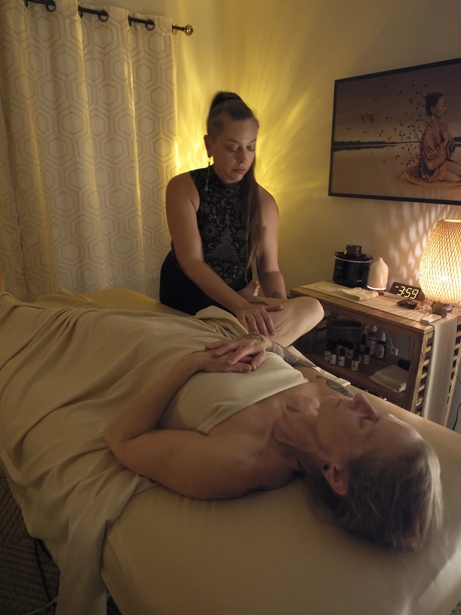 A massage table covered with a white sheet in a room with soft lighting, decorative art on the walls, a small wooden shelf with candles and bottles, a tall lamp on the right, and a patterned rug on the floor.