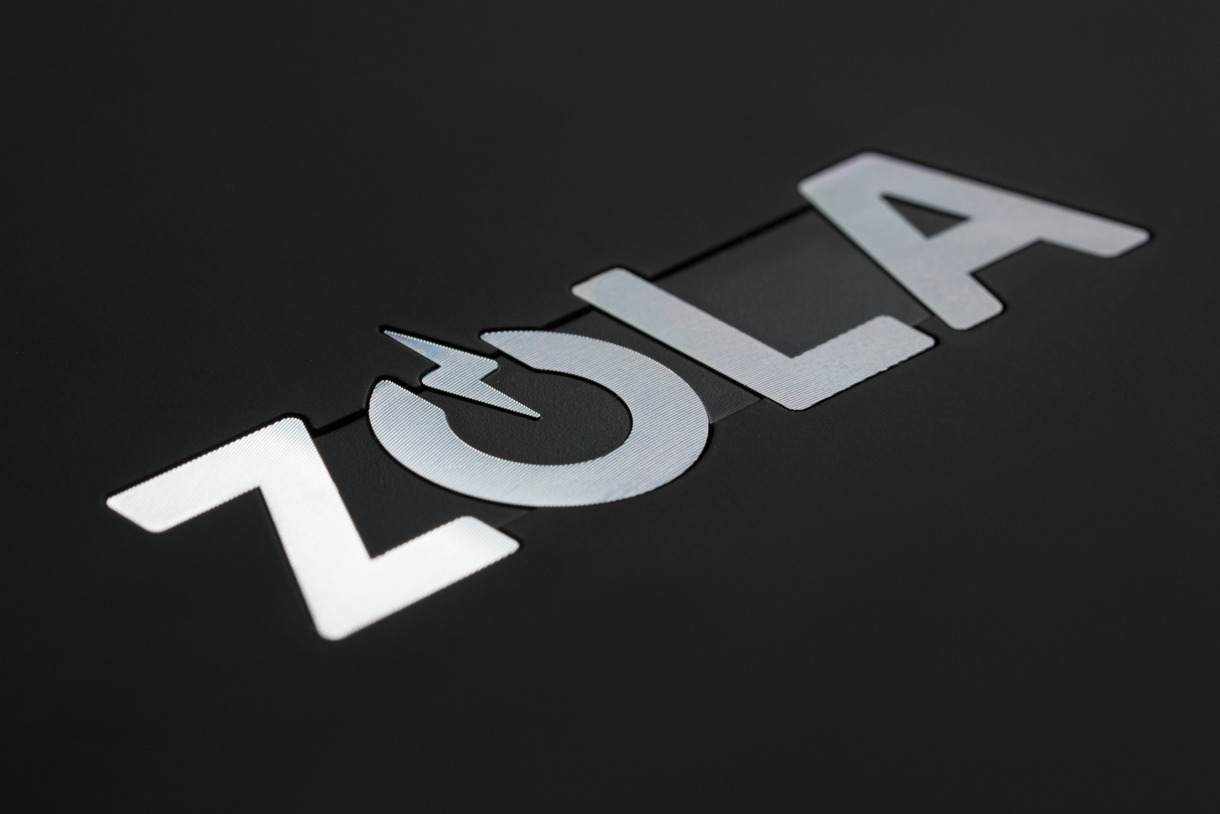 Metallic "ZOLA" logo on a dark background, featuring a power symbol as the "O."
