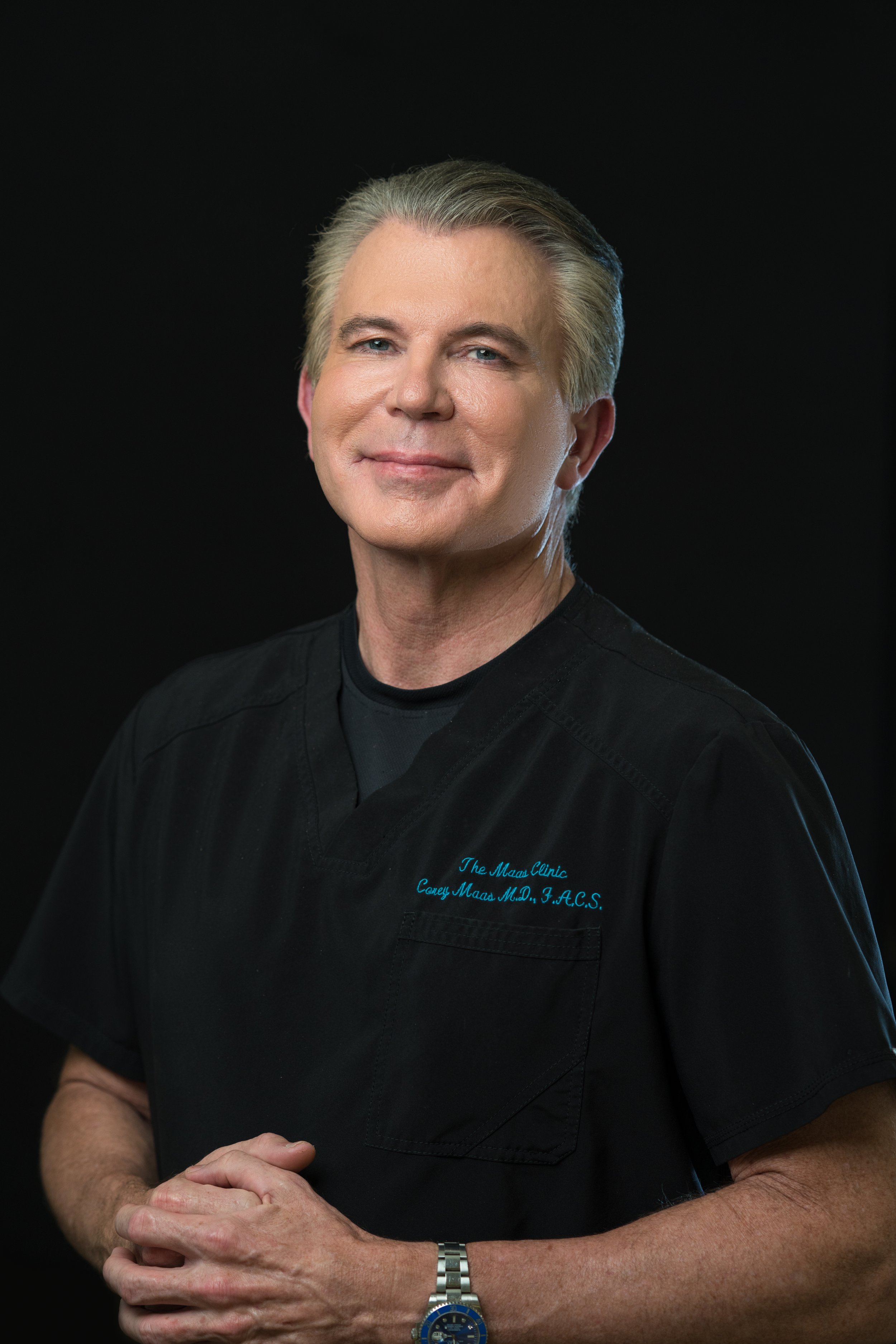 Man in black medical scrubs with text "The Maas Clinic, Casey Maas M.D., F.A.C.S." on chest, against a dark background.