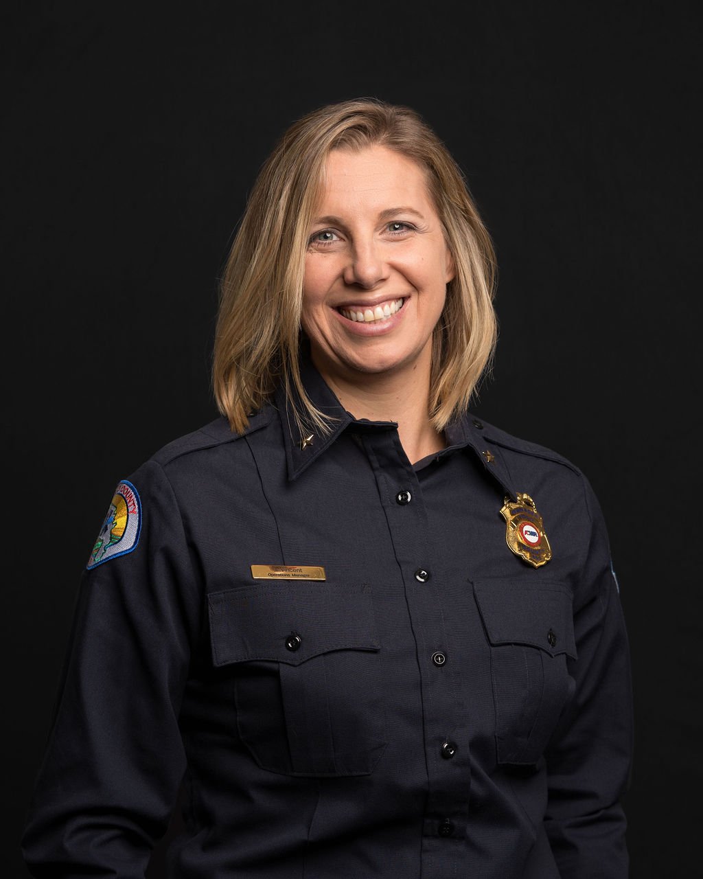 Smiling woman in uniform with a badge, against a dark background.