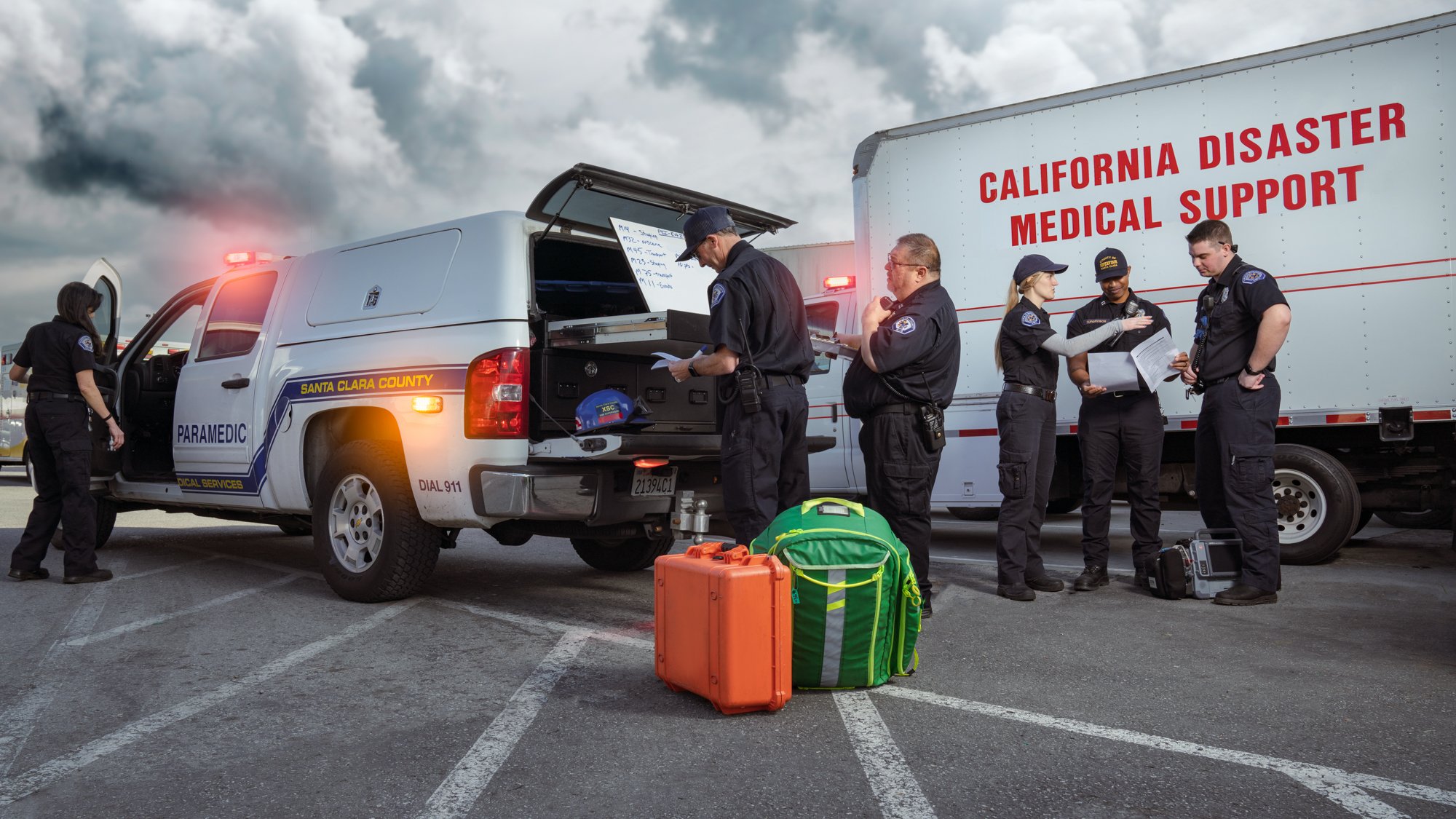 Emergency responders gathered around a Santa Clara County paramedic vehicle with equipment and a disaster support truck in the background.